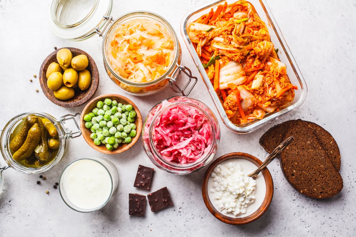 Assorted fermented foods for healthy skin displayed on a light surface, including kimchi, sauerkraut, olives, pickles, green peas, kefir, and chocolate.