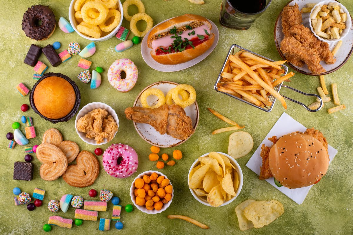 An assortment of junk food items, including fried chicken, candy, doughnuts, soft drinks, and fries, spread out on a surface not suitable for those seeking foods for healthy skin.