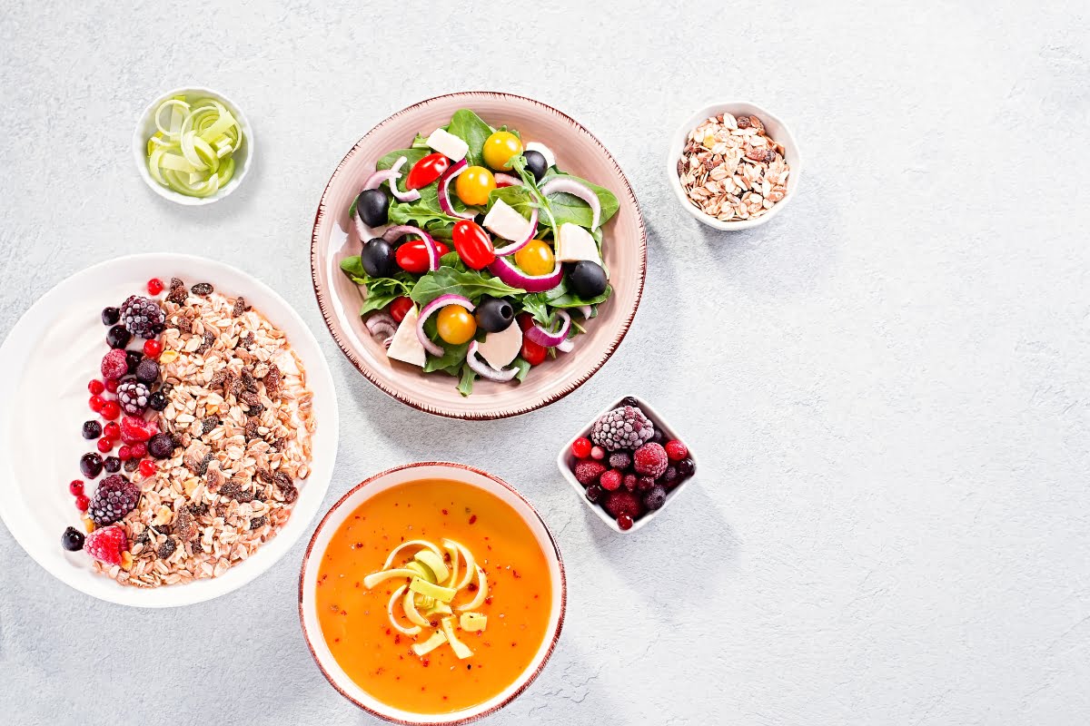 A healthy meal layout for radiant skin with a bowl of salad, a side of granola with berries, and a bowl of soup on a light-colored surface.