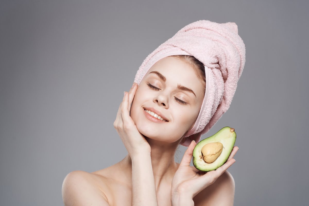 A woman with a towel on her head gently touches her face while holding a half of an avocado, a food for healthy skin.