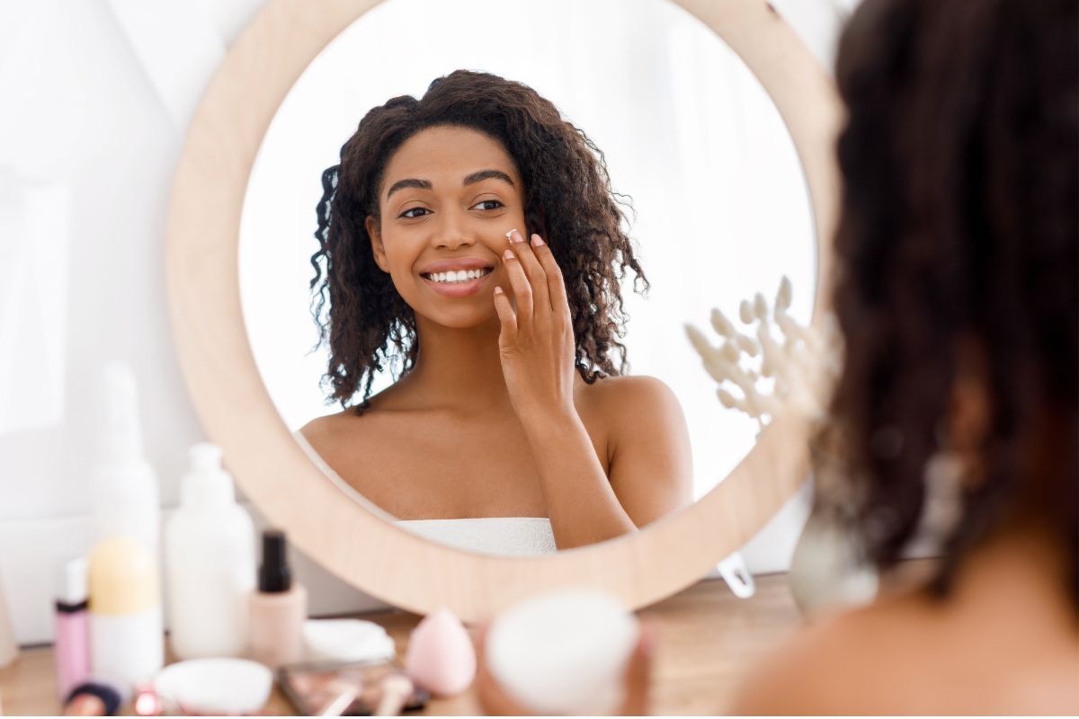 A woman applying skin regeneration cream while smiling at her reflection in a circular mirror, surrounded by beauty products.