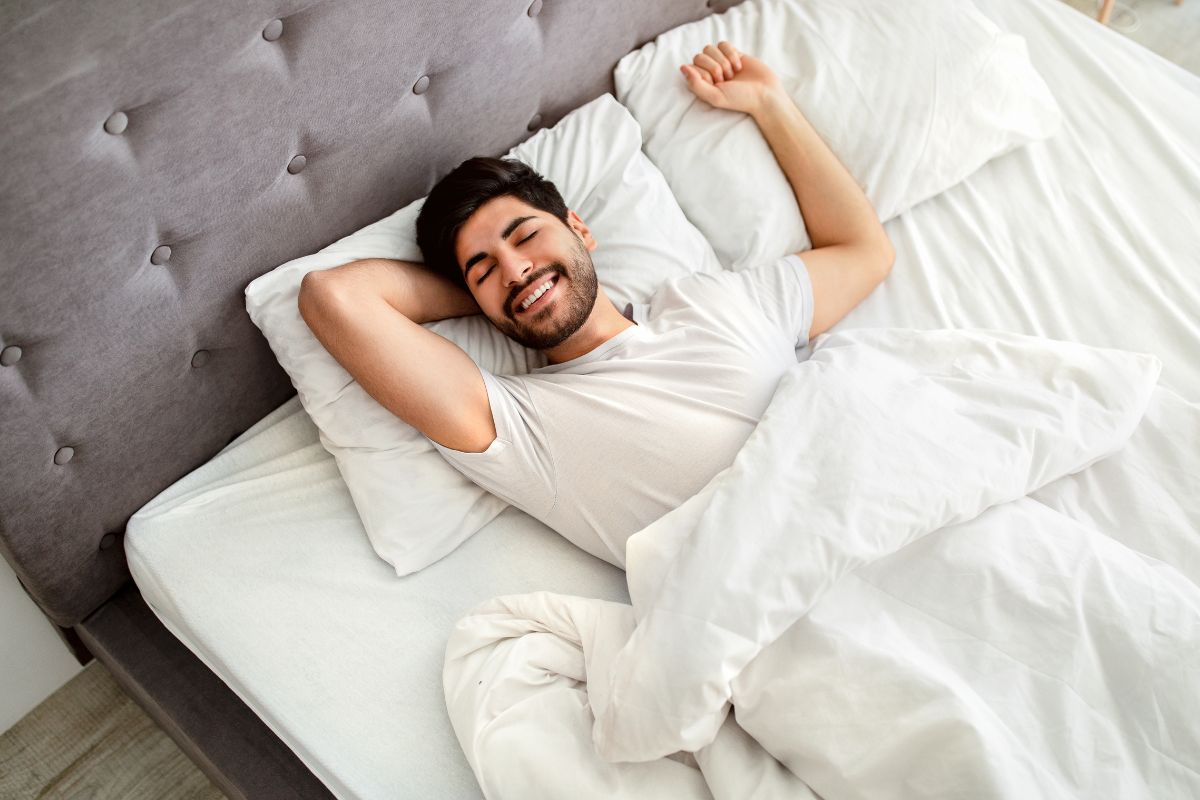 A young man smiling while stretching in bed, experiencing skin regeneration, surrounded by white sheets and a gray headboard.