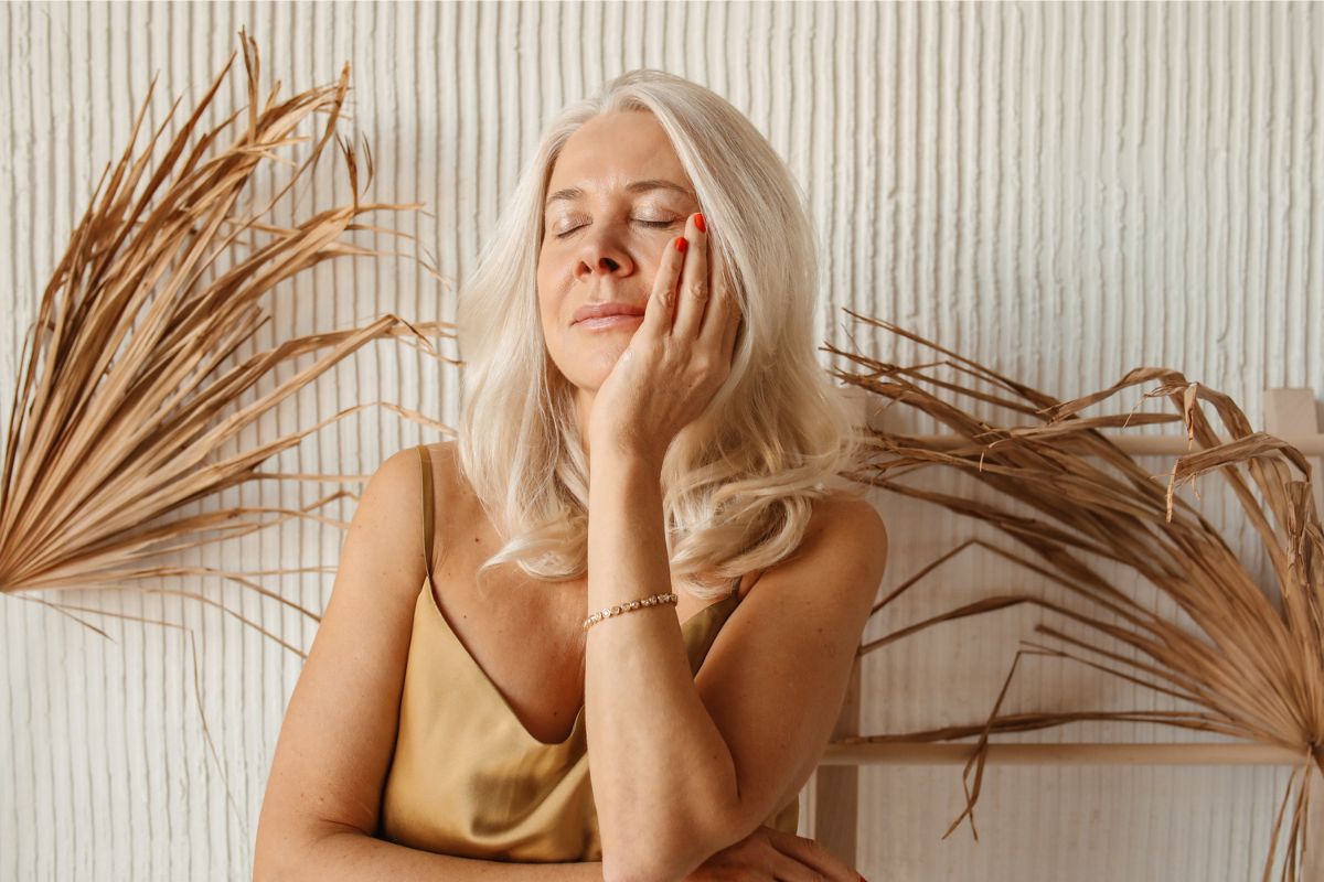 A woman with blond hair and a gold top gently touches her face, eyes closed, focusing on skin regeneration, with dried palm leaves in the background.