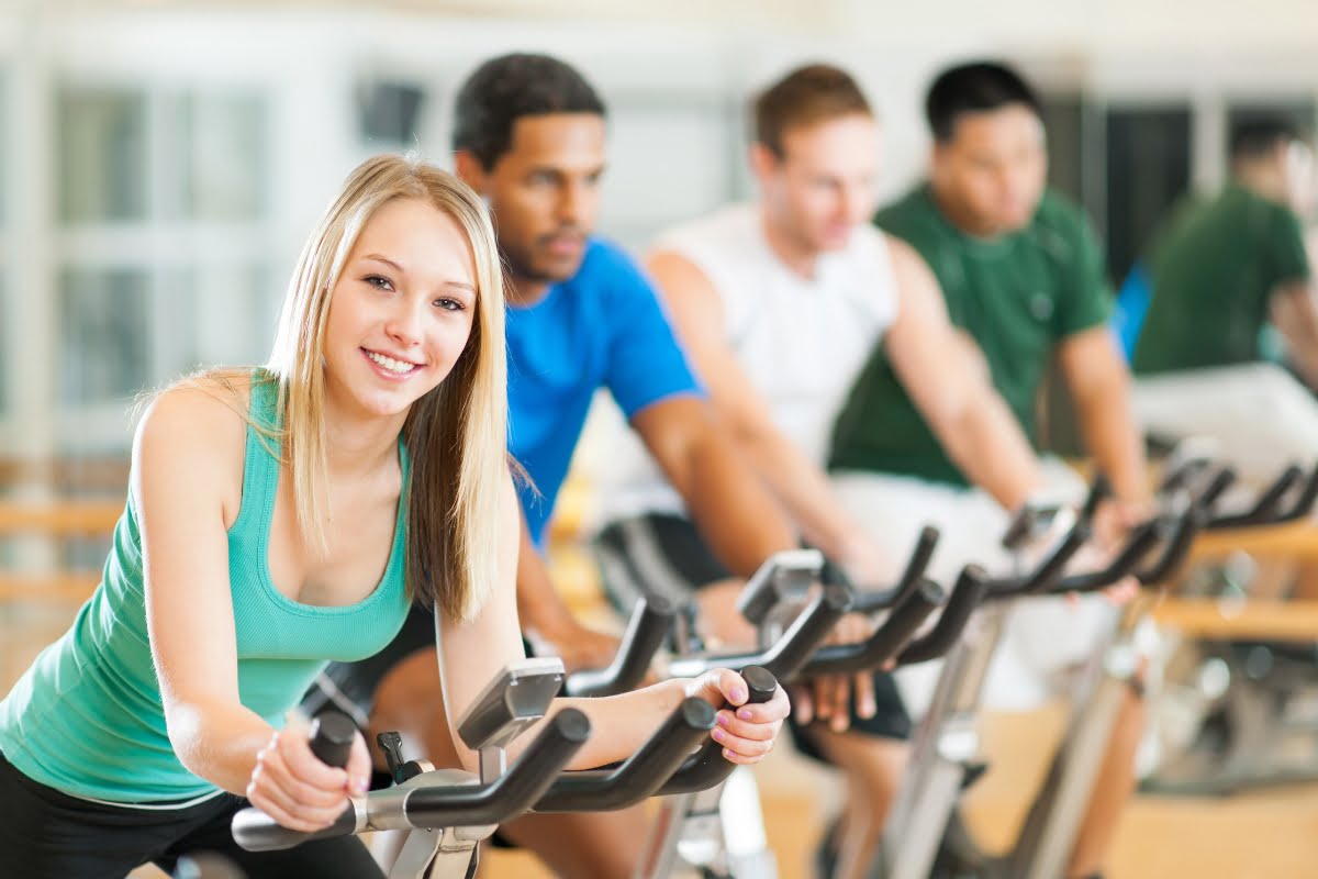 Diverse group of people enjoying the exercise benefits on stationary bikes in a gym, smiling and looking towards the camera.