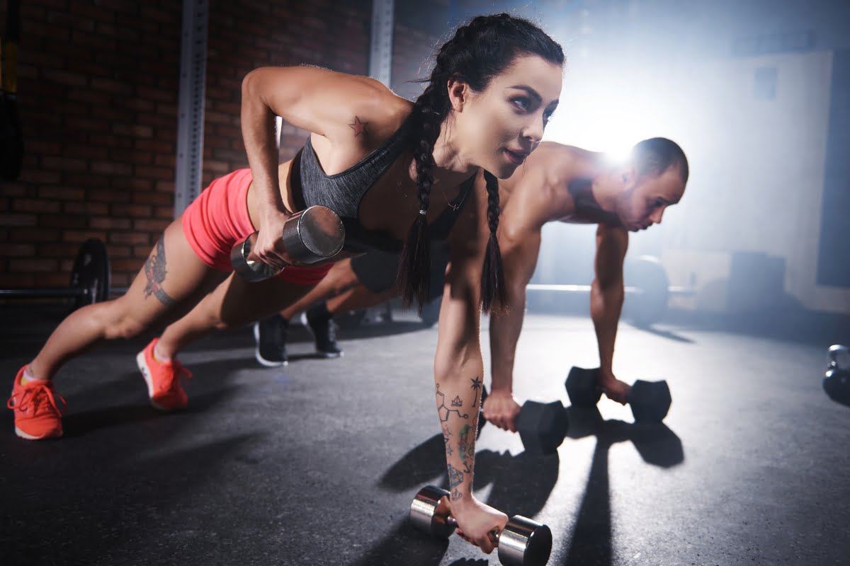 Two people performing plank exercises with dumbbells in a gym, focusing intently on the exercise benefits of their workout.