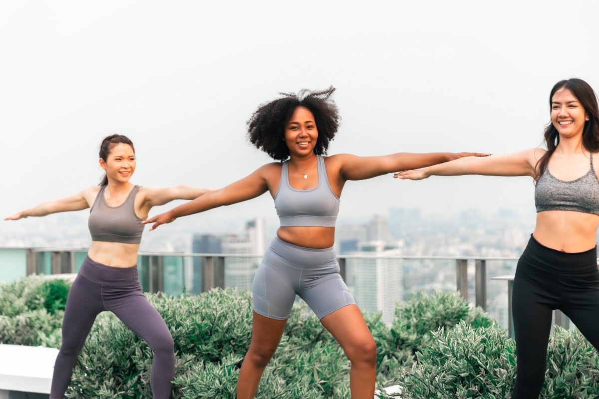 Three women in athletic wear demonstrating the exercise benefits of yoga poses together on a rooftop with a cityscape in the background.