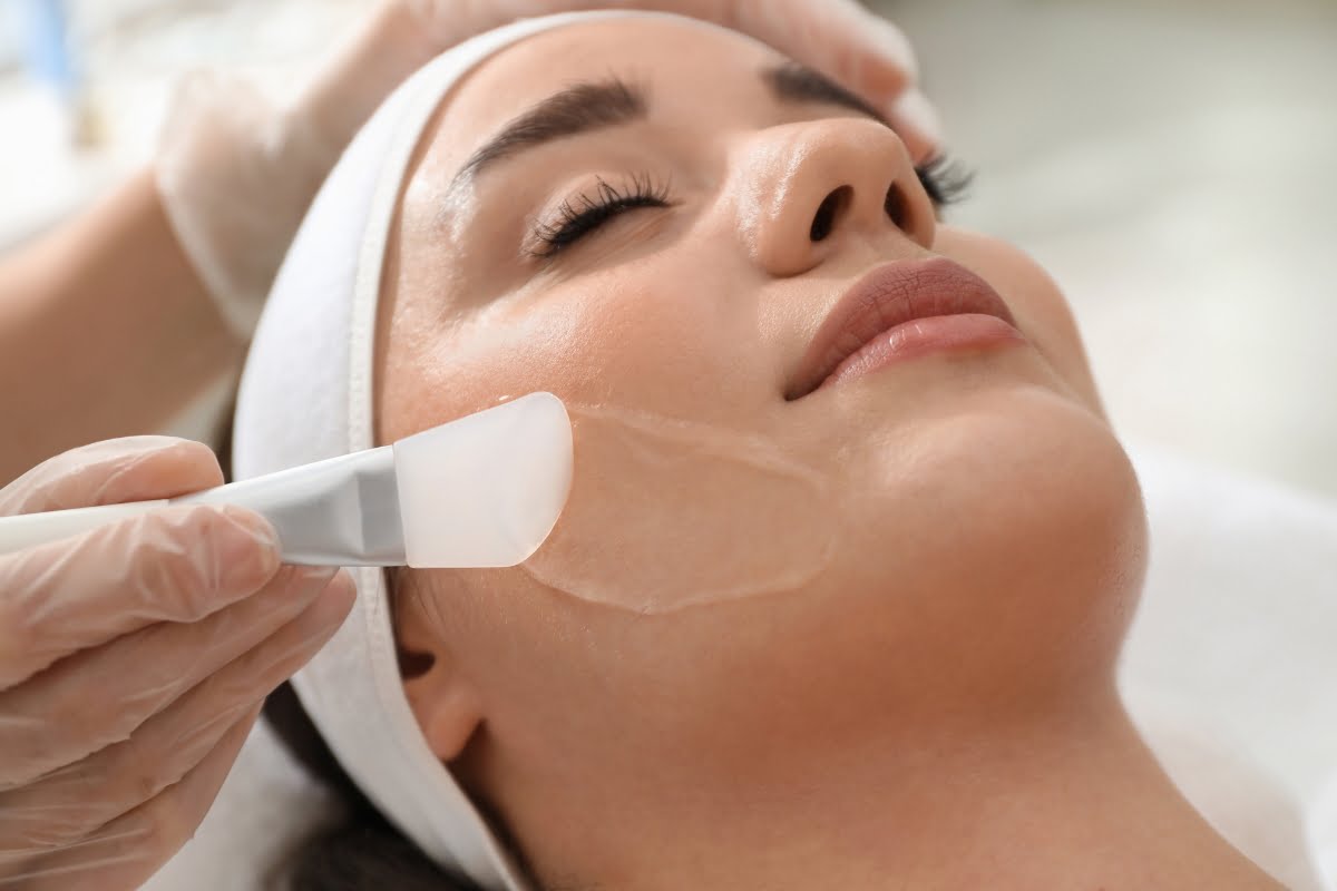 Aesthetician conducting physical exfoliation by applying a treatment mask on a woman's cheek using a brush in a spa setting.