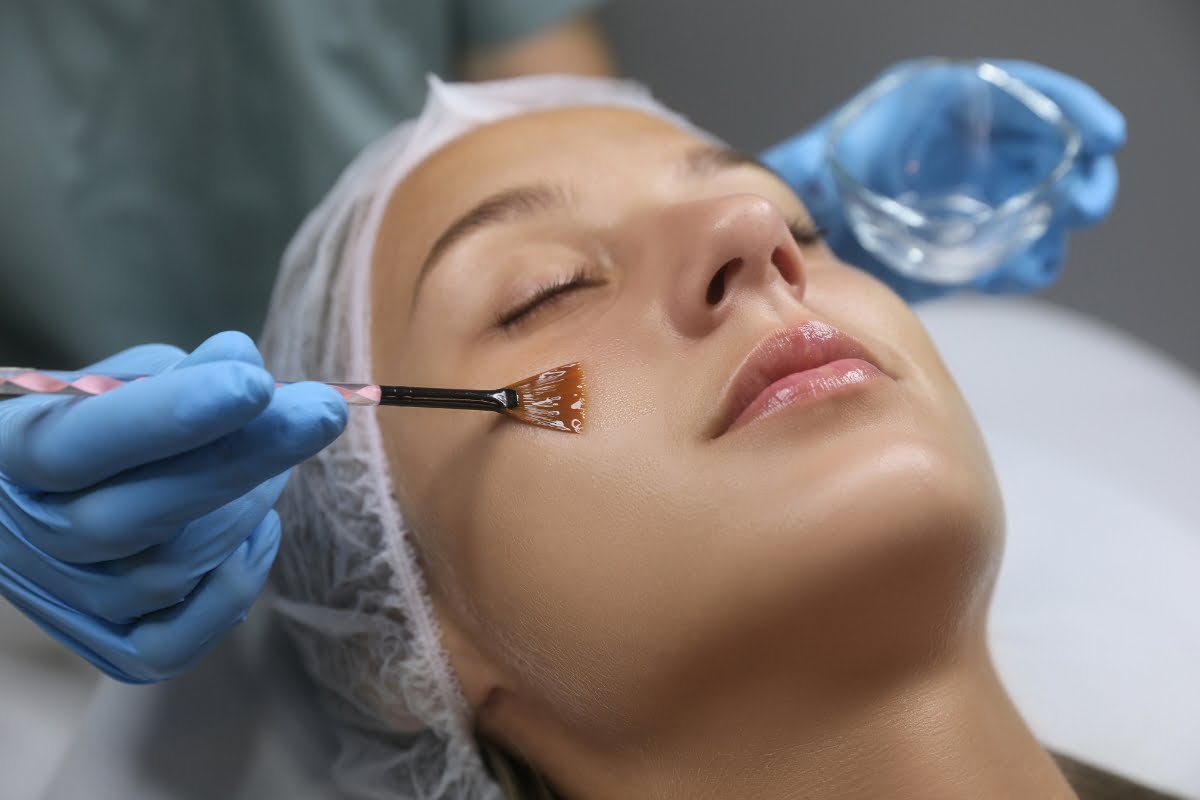 A woman receives a facial treatment, with a professional applying physical exfoliation to her cheek using a brush.