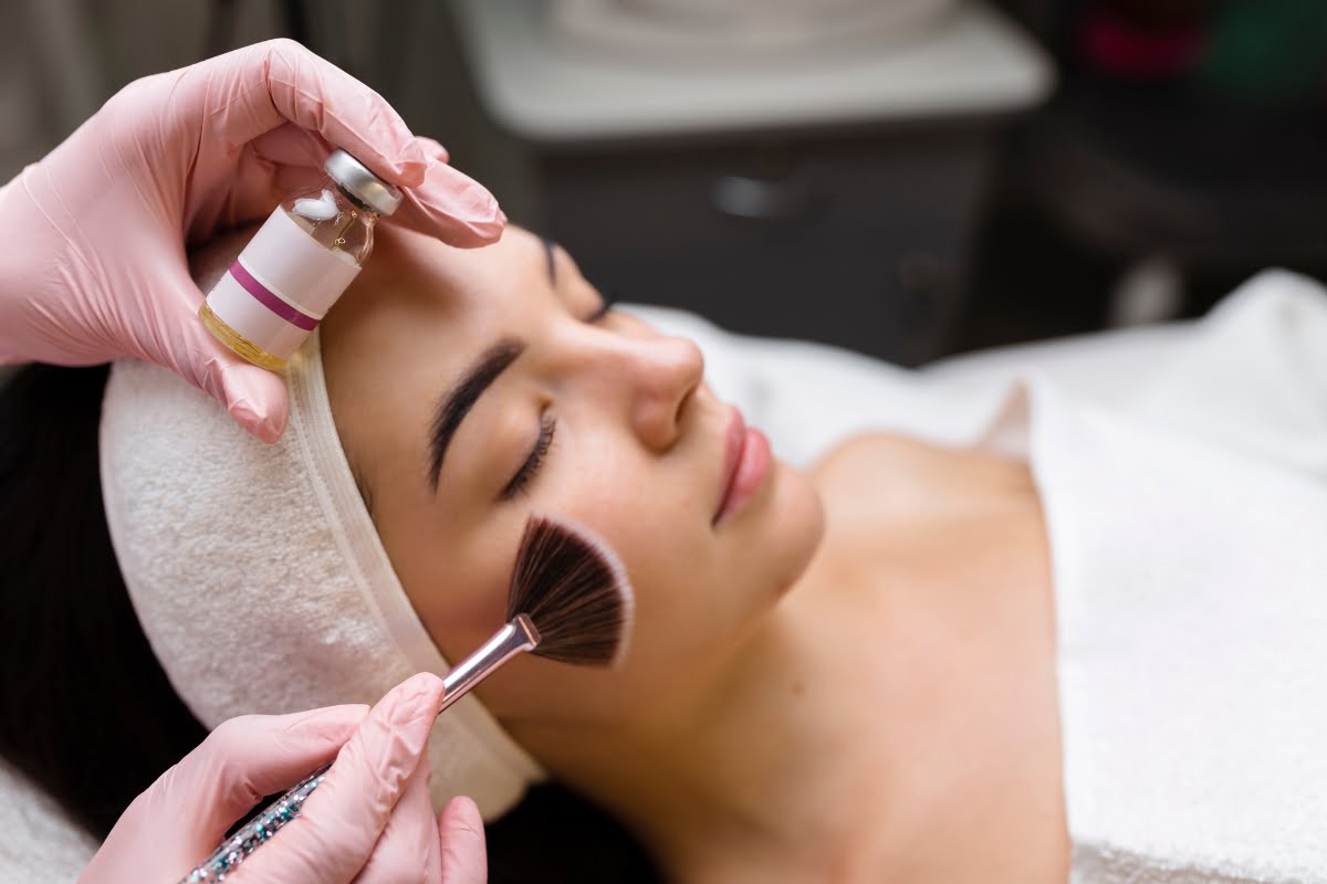 A woman receiving a physical exfoliation treatment with a brush applying a serum on her cheek in a serene spa setting.