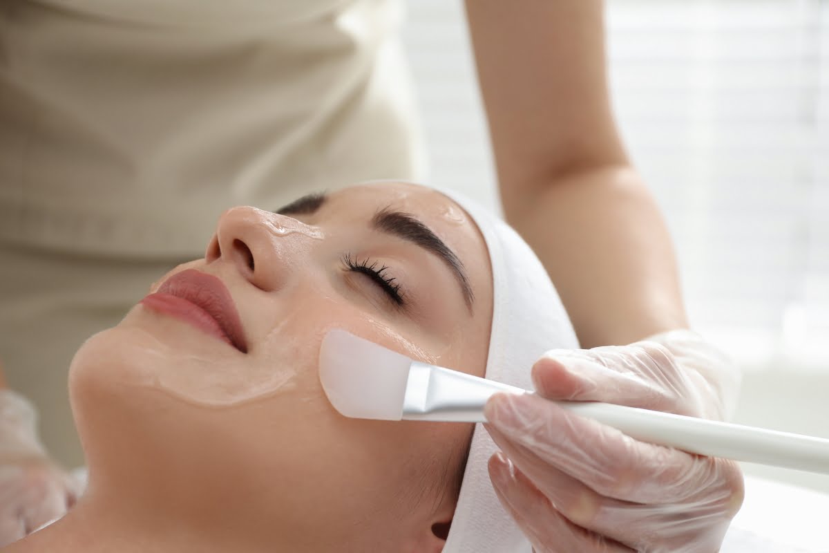 A woman receiving a physical exfoliation treatment with a mask being applied by a professional using a brush.