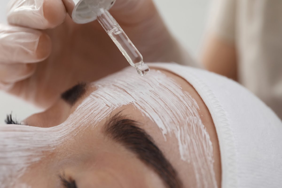 Close-up of a person receiving a physical exfoliation treatment with a serum being applied on their forehead covered in a white mask.