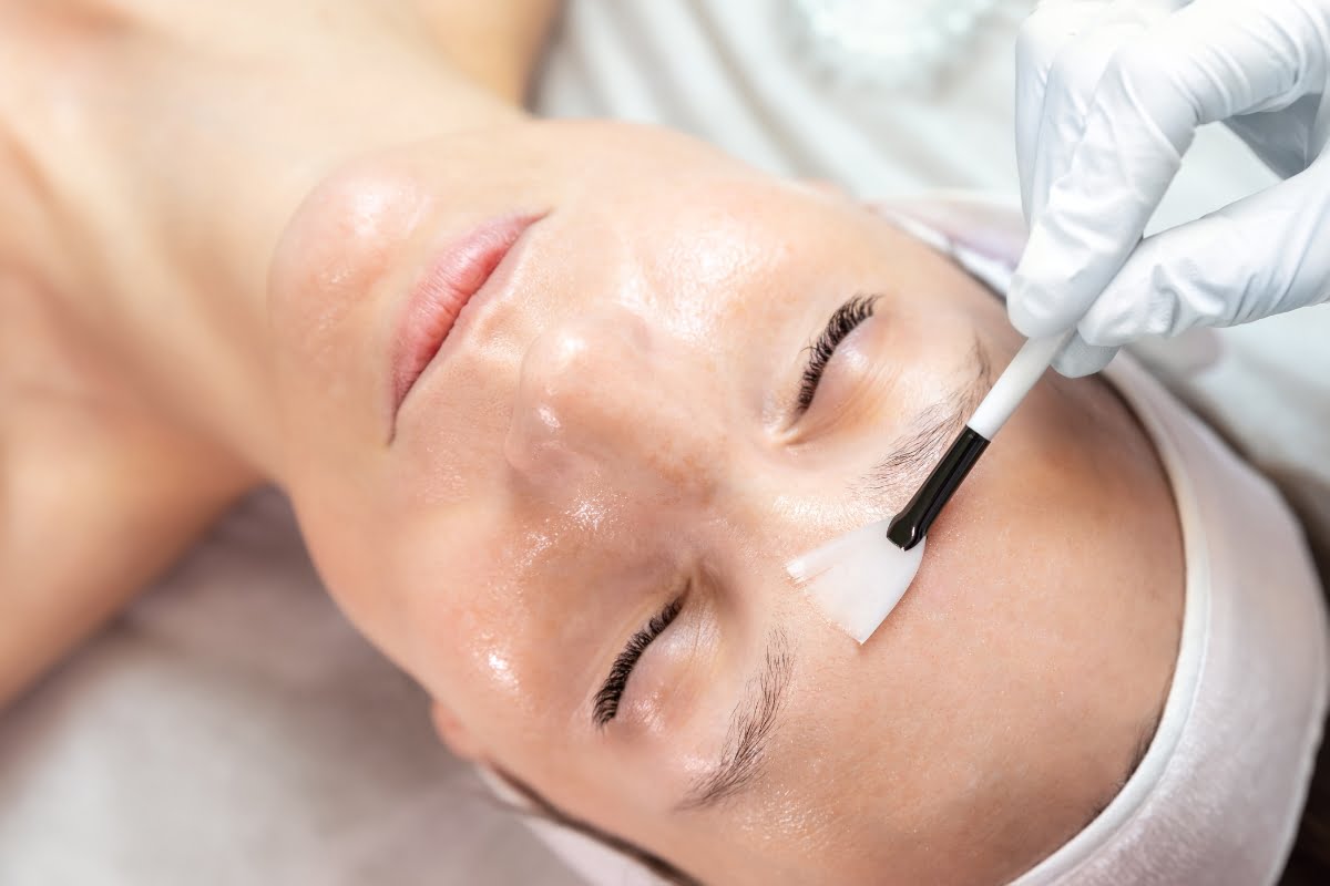 A woman is receiving an eyelash treatment, with an esthetician performing physical exfoliation and applying a solution to her lashes using a small brush.