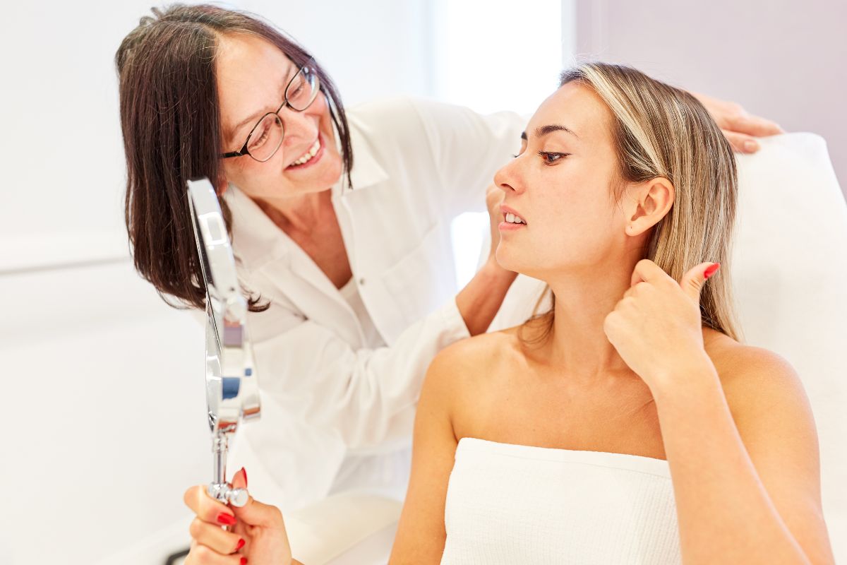 A woman examines her face in a hand mirror while another woman, wearing glasses and a white coat, smiles and looks on supportively, potentially answering the question, "What is mesotherapy?