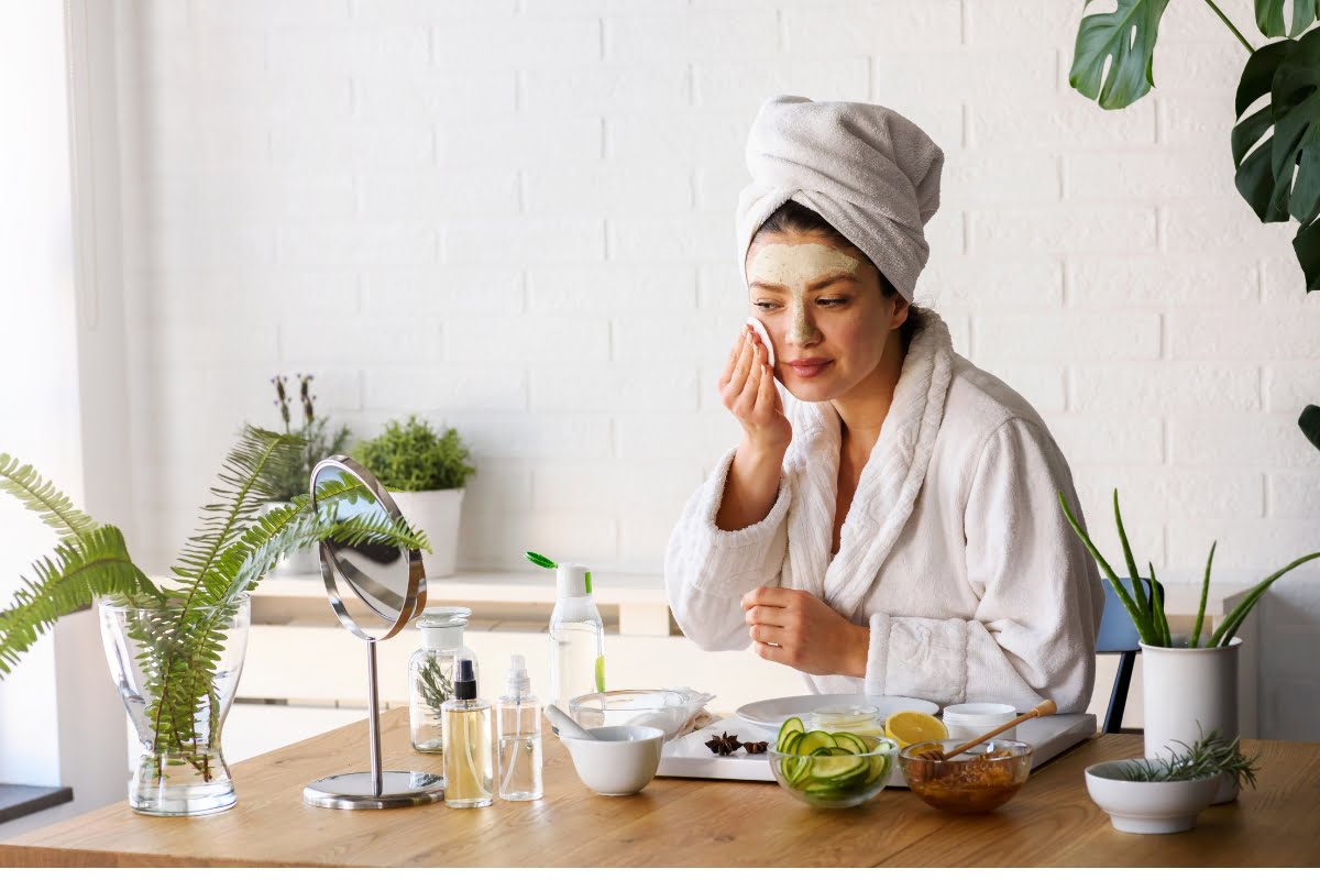 A woman in a white bathrobe and towel turban applies a facial mask while sitting at a table with skincare products and plants, seamlessly blending her skin care routine with stress management.