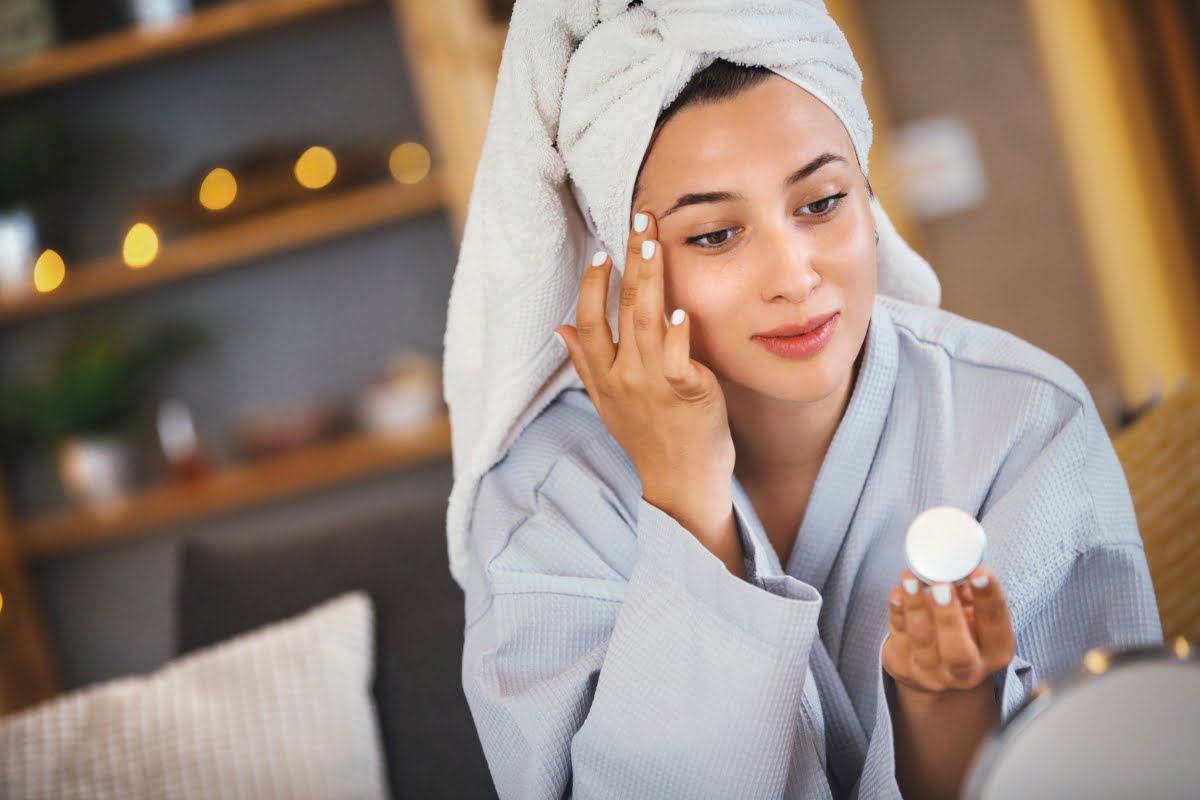 A person with a towel on their head and wearing a robe applies cream to their face while looking into a mirror, incorporating stress management into their skin care routine.