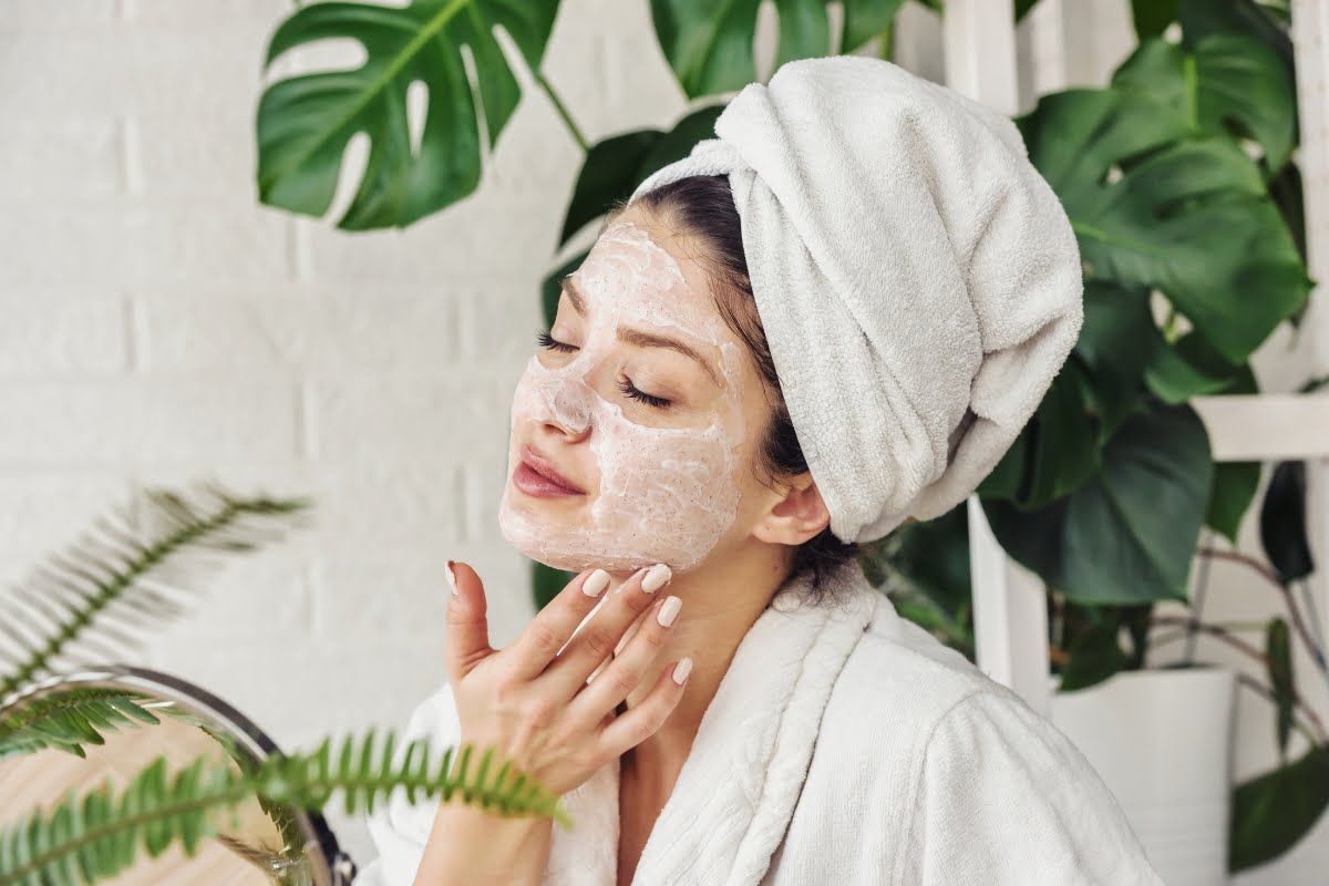 A person in a bathrobe and towel turban applies a face mask, enjoying their skin care routine amidst the calming presence of potted plants.