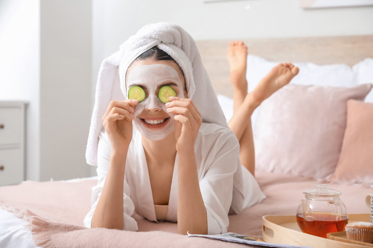 A person lies on a bed with a facial mask on and a towel wrapped around their head, holding cucumber slices over their eyes. Engaged in their skin care routine, a tray with a teacup and jar rests beside them, serving as part of their stress management ritual.