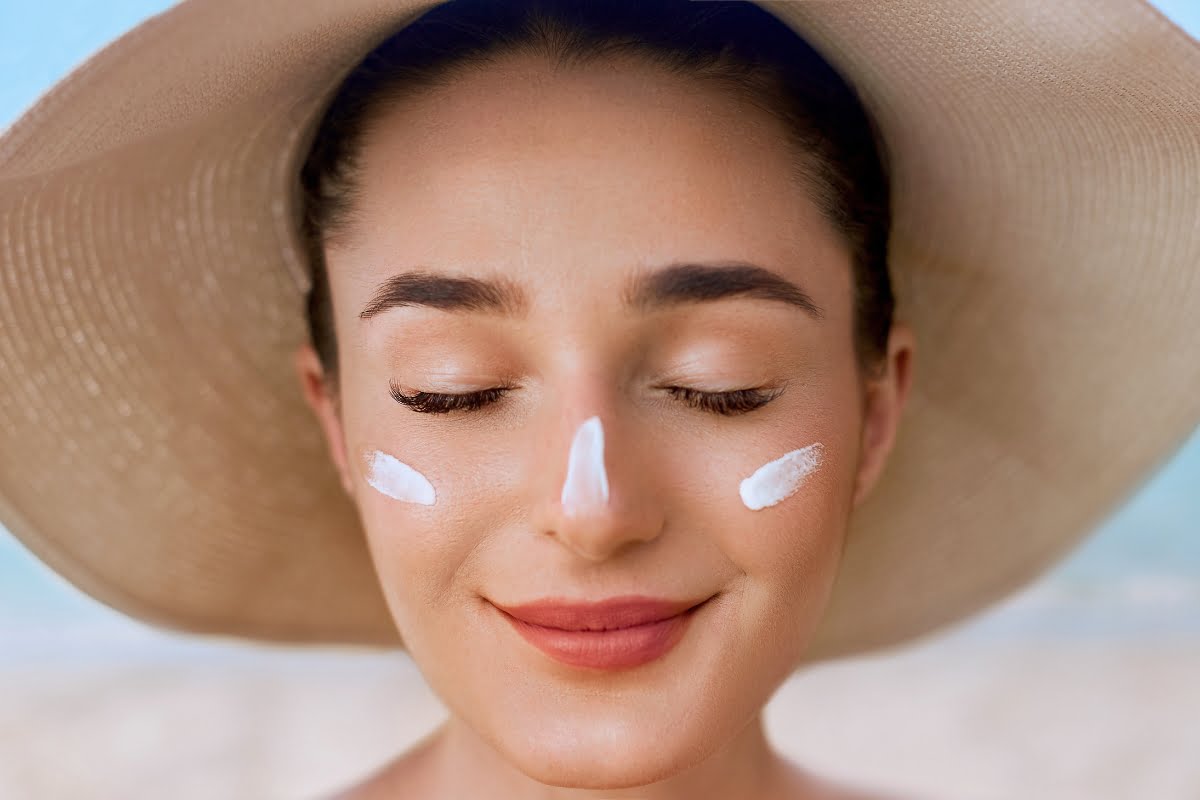 Woman with closed eyes and slight smile, wearing a wide-brimmed hat and sunscreen on her nose and cheeks, partaking in her skin care routine while standing outdoors.