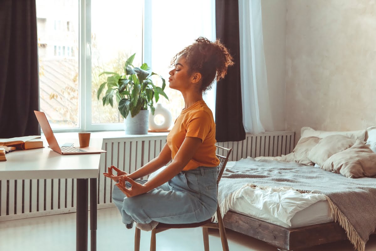 Woman in orange shirt and blue jeans sitting on a chair in a bedroom near a desk, meditating with eyes closed, hands in a mudra gesture, laptop and plants nearby. This serene moment is part of her stress management routine.