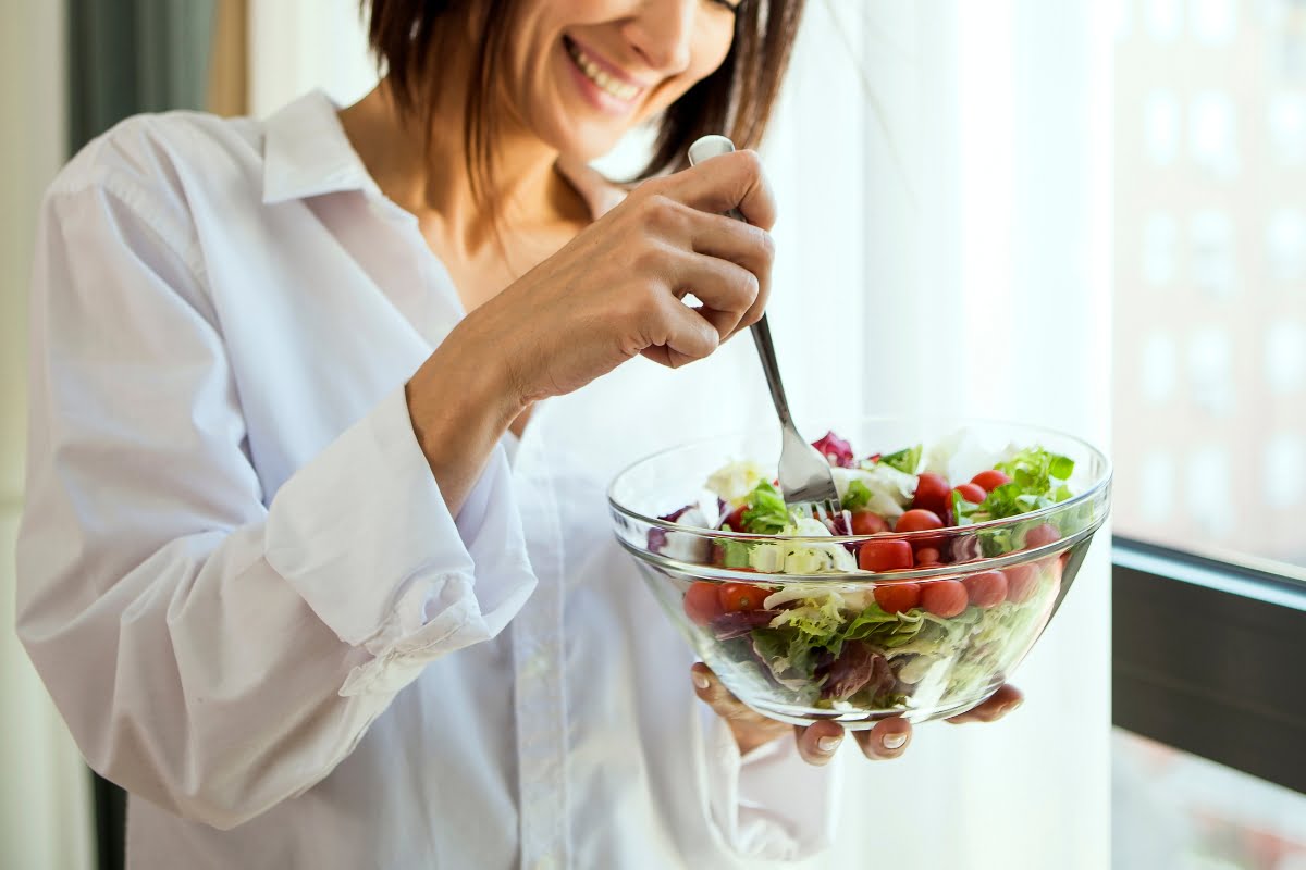 A person in a white shirt smiles while holding a glass bowl of mixed salad with cherry tomatoes, near a window, as part of their stress management routine.