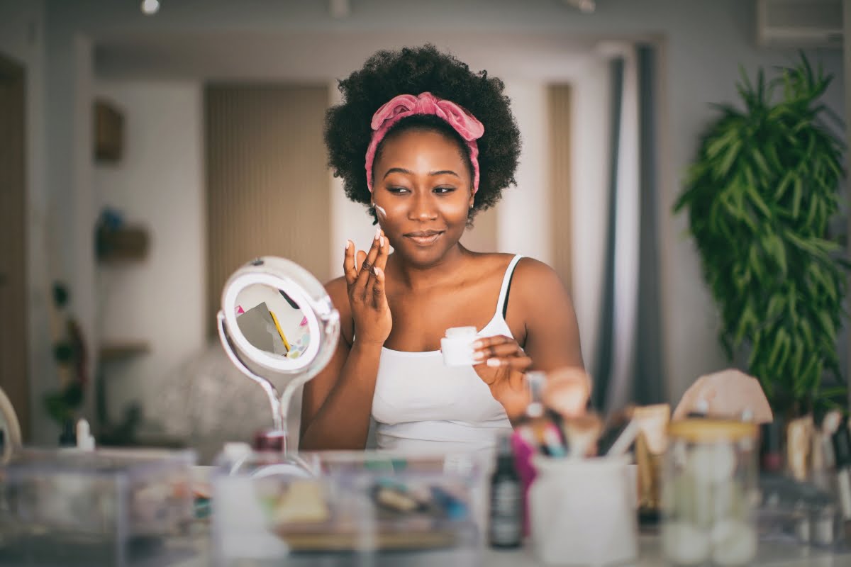 A woman with a pink headband applies skincare from a small jar while smiling in front of a vanity mirror, seamlessly integrating her skin care routine into her stress management. Various cosmetic items are scattered on the surface.