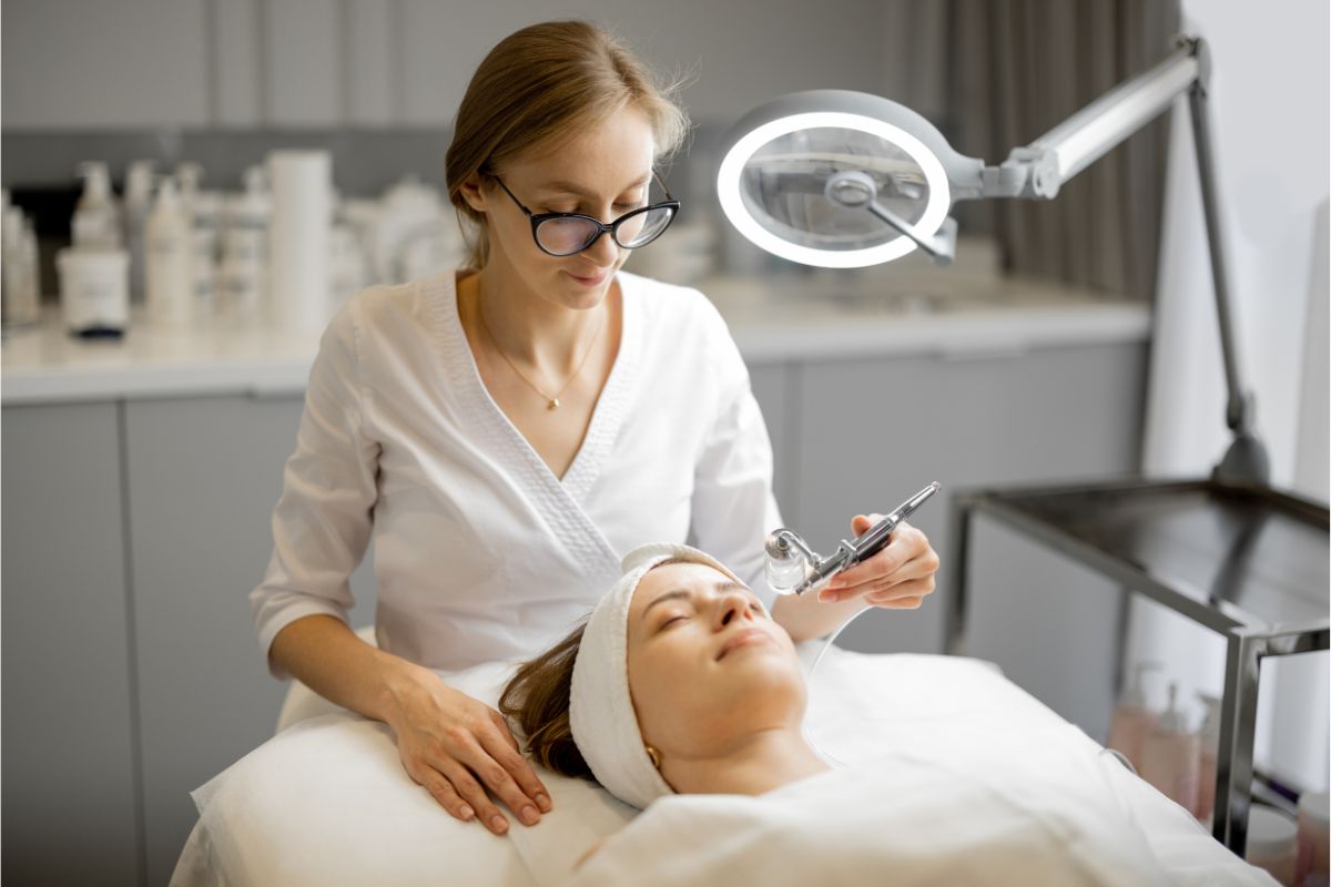 A beautician wearing glasses performs a facial treatment on a reclining client under a bright lamp in a professional skincare clinic, answering the client's questions about what is mesotherapy.