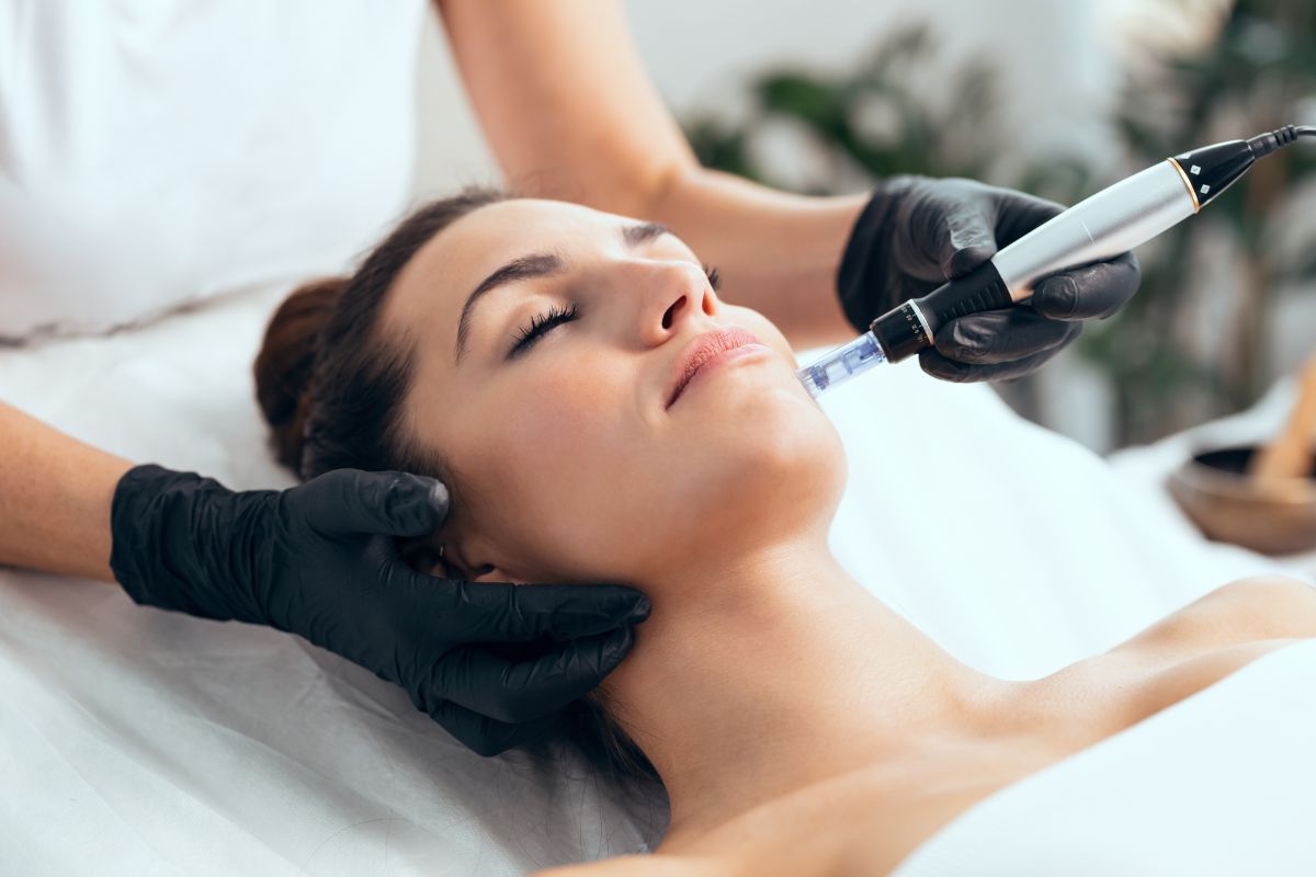 A woman lying on a treatment bed receives a facial procedure from a professional wearing black gloves, who is using a small tool on her face—could this be the answer to what is mesotherapy?