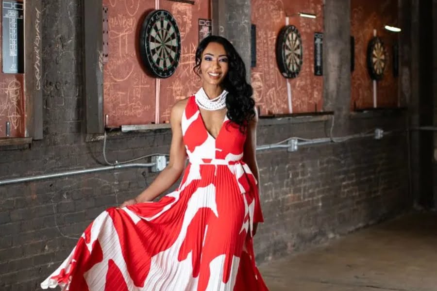 Dr. Yolanda Scott, wearing a red and white dress, stands in front of dartboards mounted on a brick wall, holding her dress and smiling.