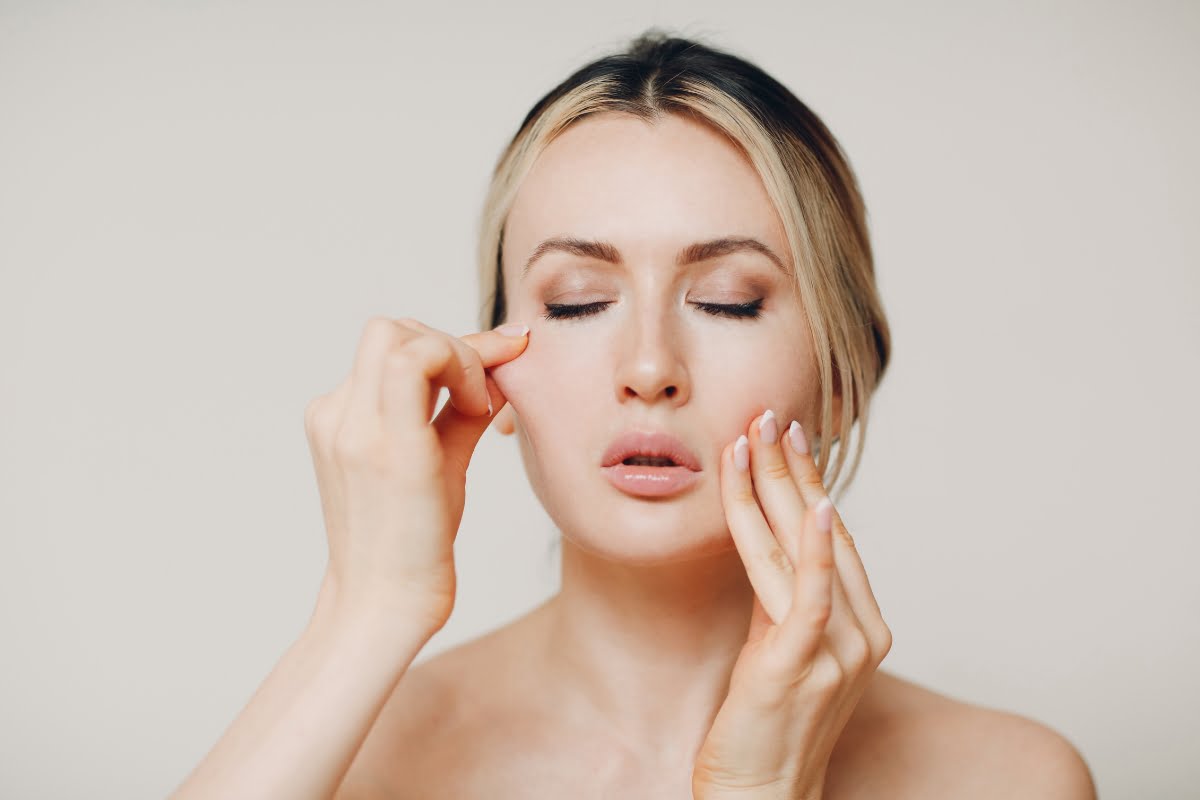 A person with closed eyes gently pinches their cheeks, appearing calm and focused, showcasing their skin elasticity against a plain background.