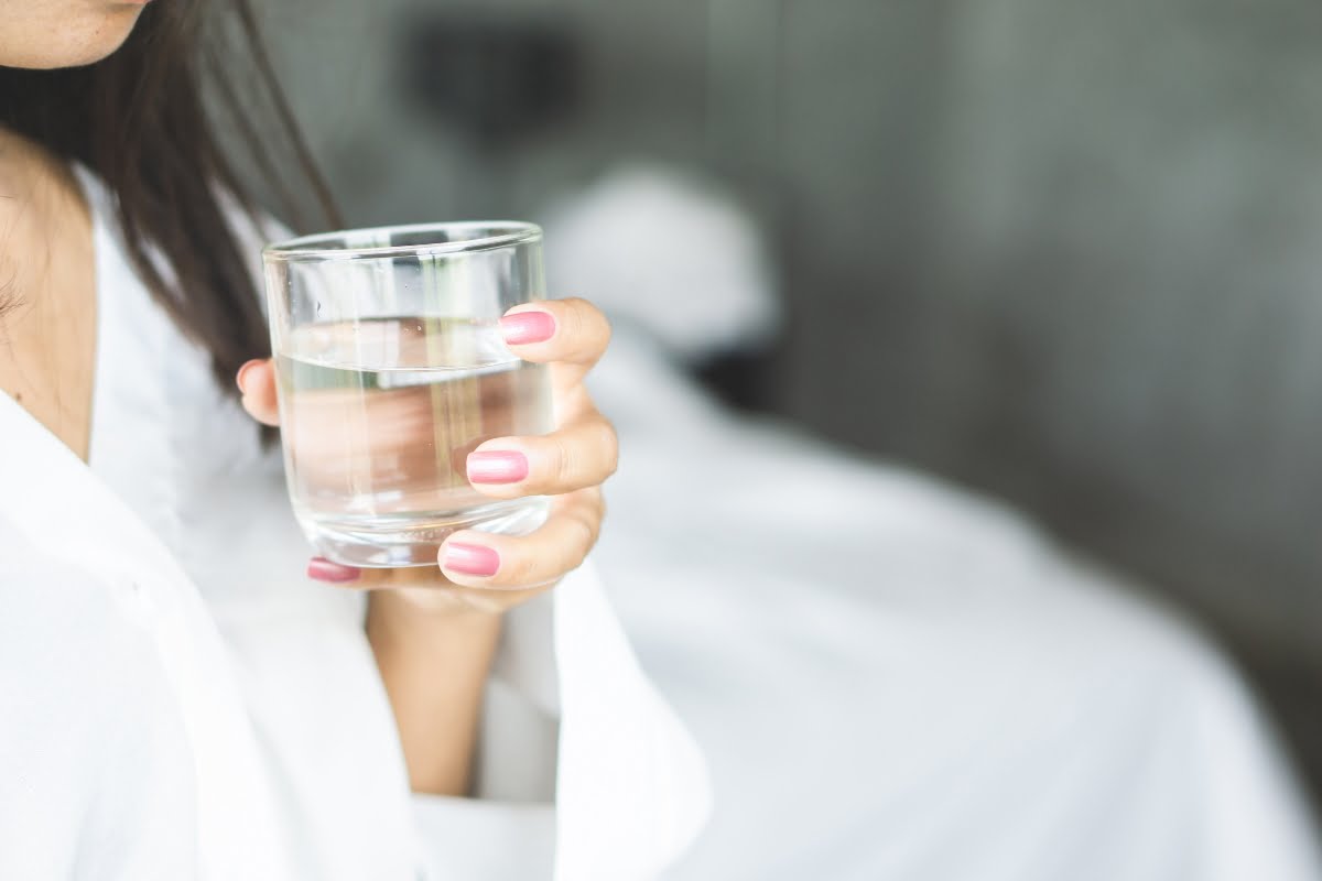 A person with pink nail polish holds a glass of water close to their mouth while wearing a white garment, showcasing their skin elasticity.