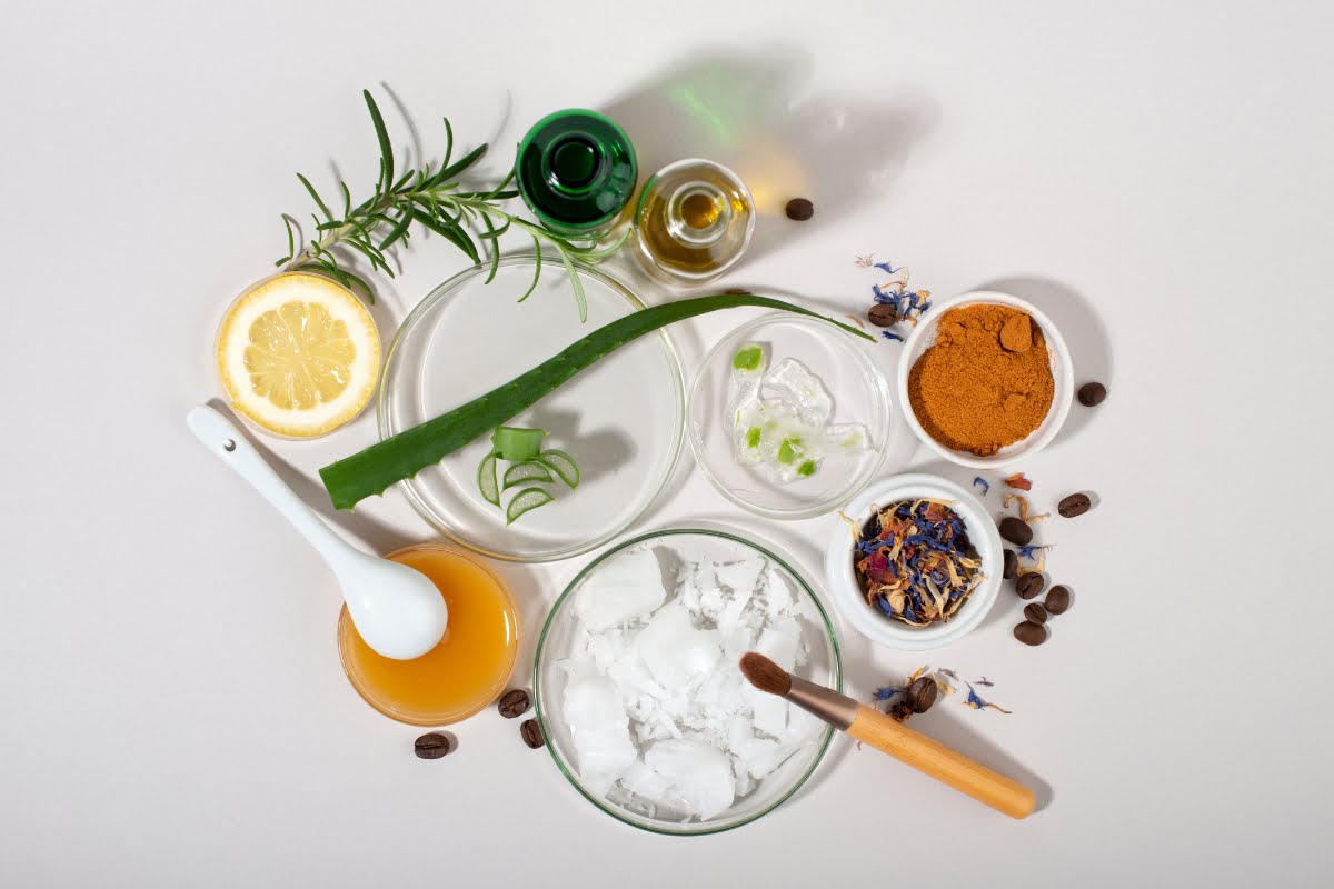 A flat lay of natural skincare ingredients on a white surface, including aloe vera, lemon, oils, honey, coconut oil, spices, and dried flowers, thoughtfully arranged around glass bowls and a spoon—perfect for boosting skin elasticity.