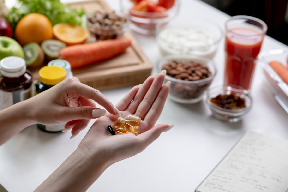 A person holds various supplement pills in one hand, with a table full of fresh fruits, vegetables, nuts, and a glass of juice in the background, all contributing to improved skin elasticity.