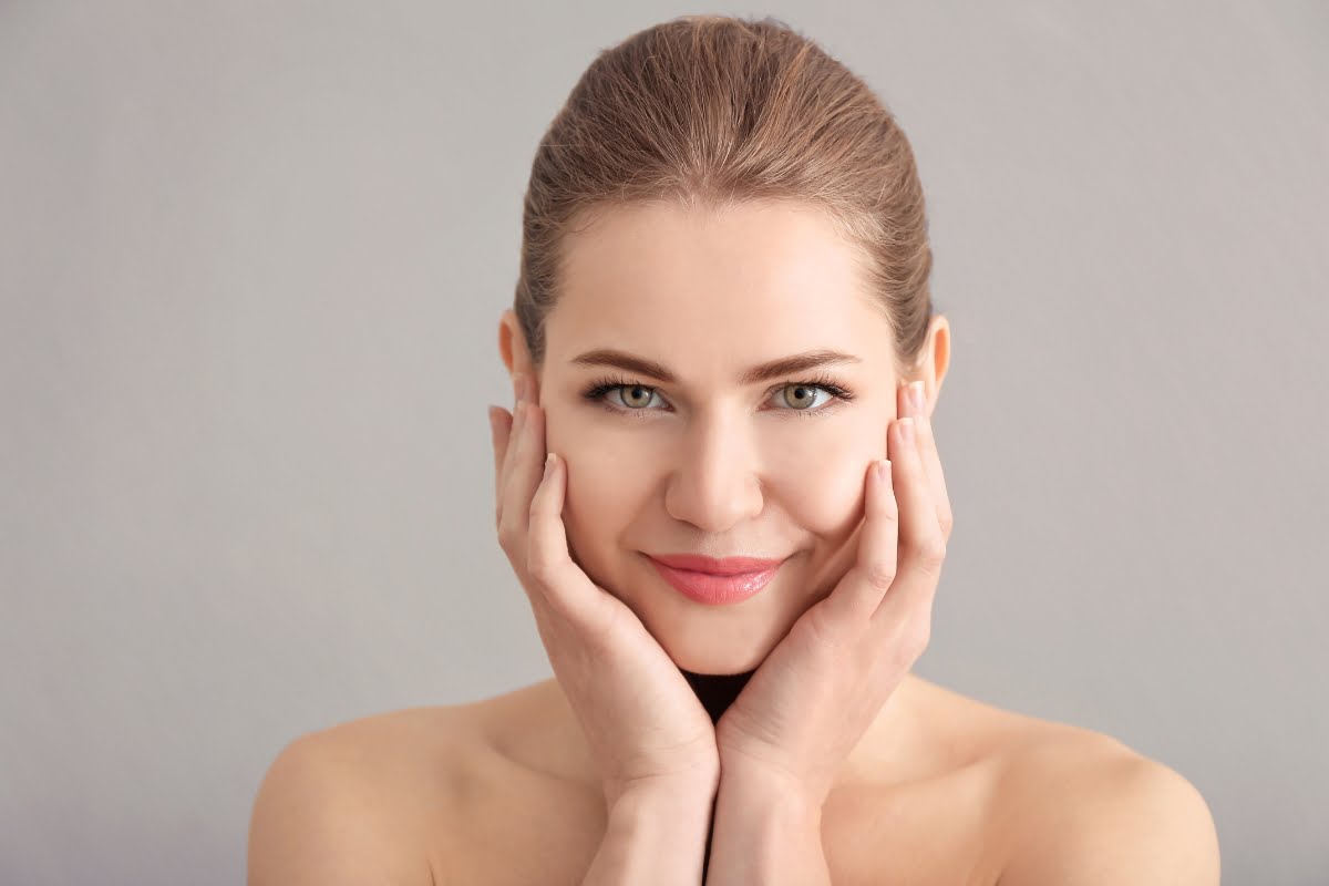 A woman with light brown hair pulled back, smiling softly, with both hands gently touching her cheeks, highlighting her skin's elasticity against a plain gray background.