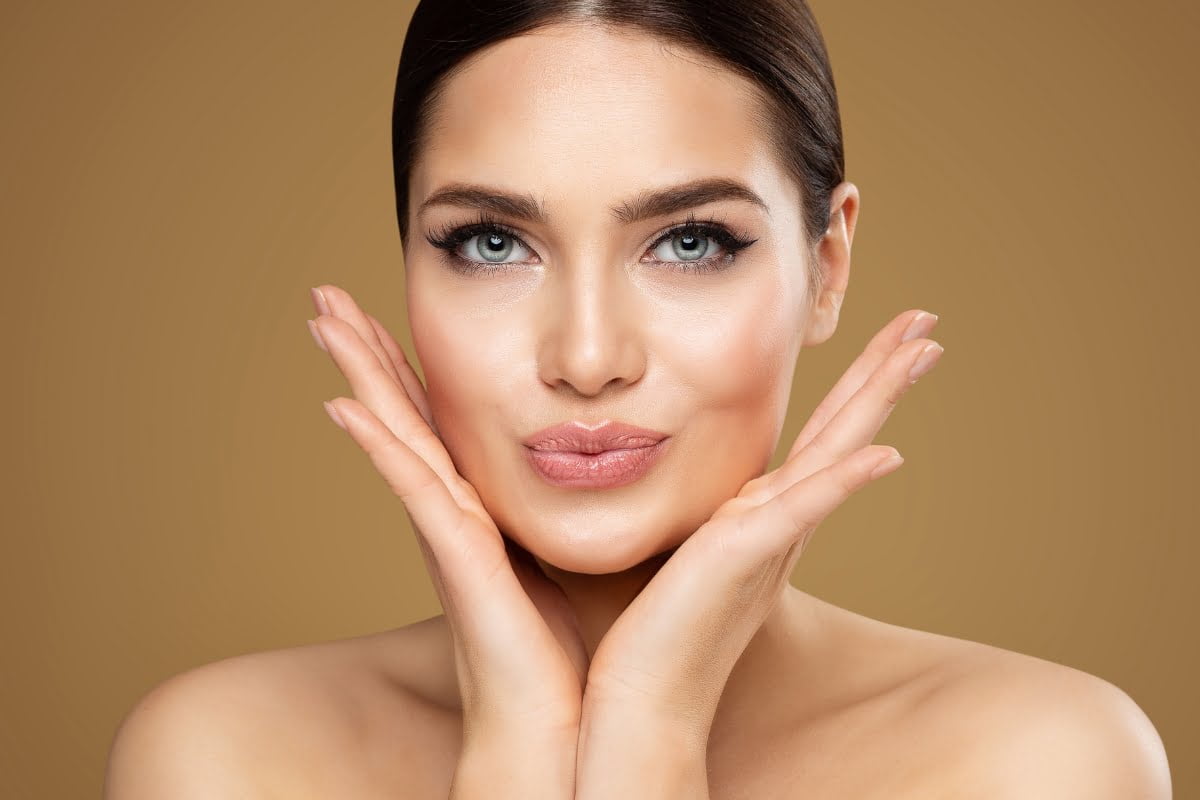 A person with smooth skin, possibly enhanced by skin fillers, and styled hair poses with hands framing their face against a brown background.