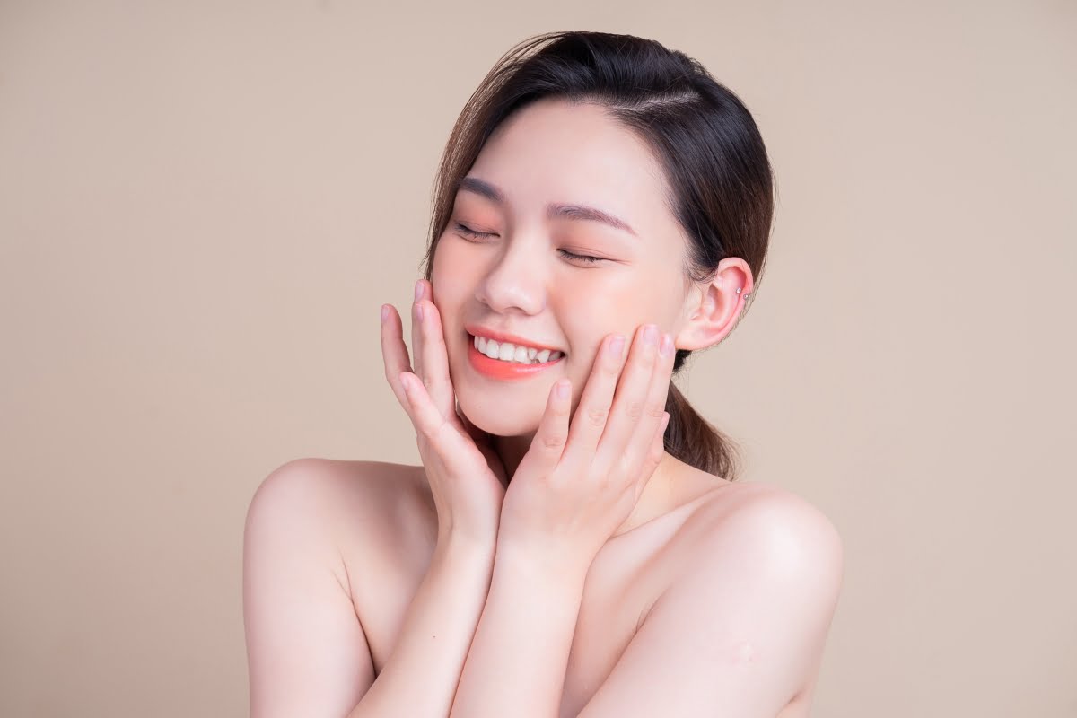 A smiling woman with closed eyes touches her face with both hands against a neutral background. Her clear skin, possibly enhanced by skin fillers, suggests contentment and relaxation.