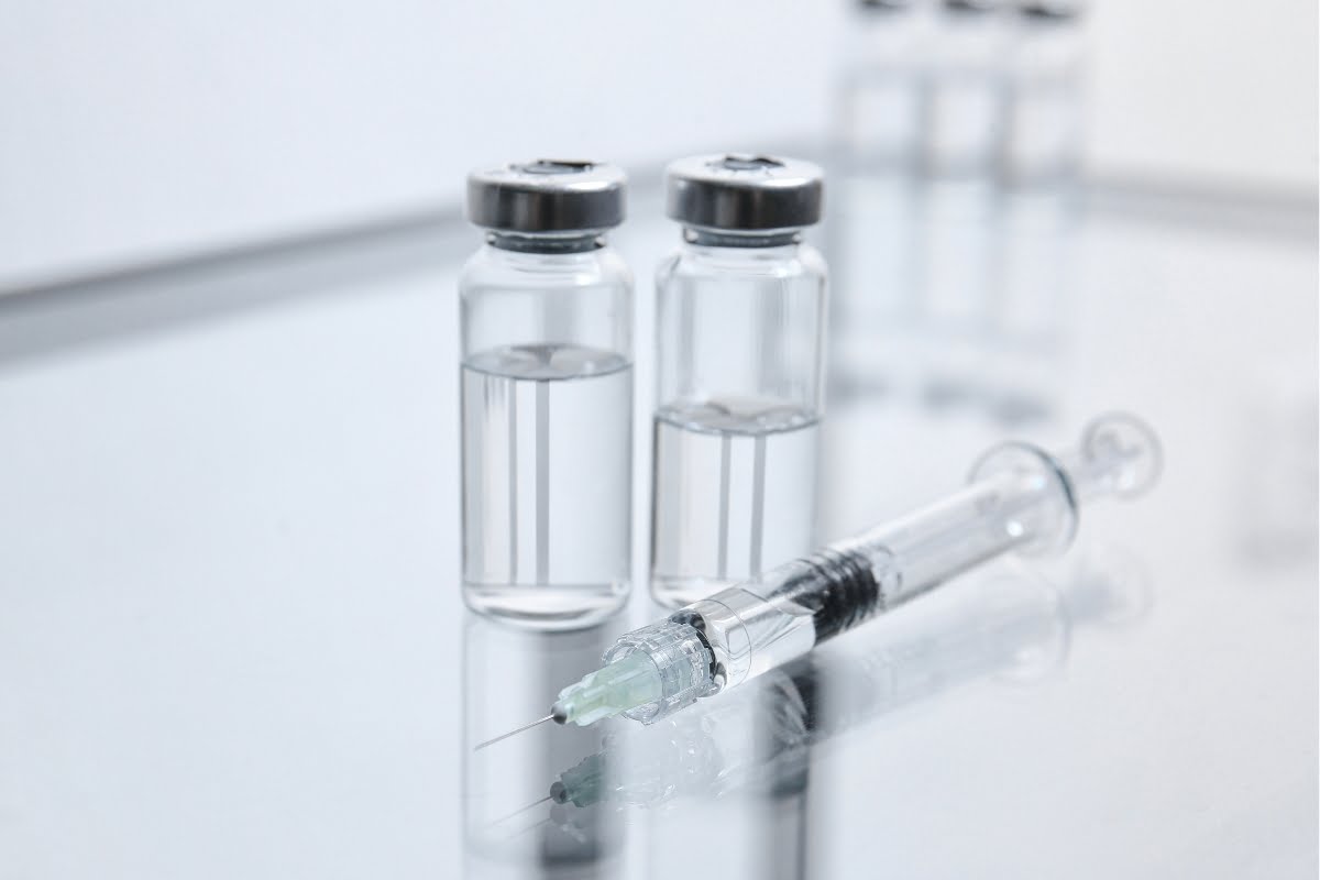 Two vials of clear liquid, custom-tailored for skin fillers, are placed on a reflective surface next to a syringe. The background is out of focus.