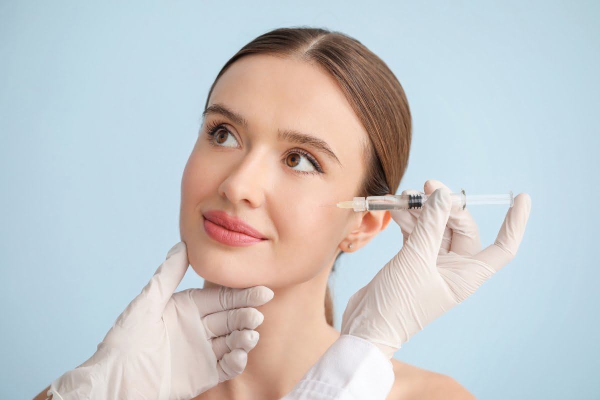 A woman with brown hair receives a skin filler injection in her cheek from a gloved professional against a light blue background.