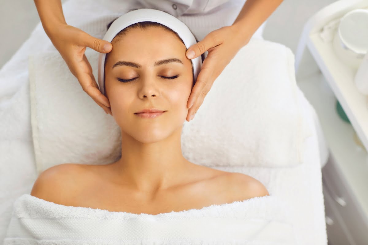 A person lies down with eyes closed, receiving a facial massage. The individual's head is wrapped in a towel, and their shoulders are covered. This relaxing treatment often precedes skin fillers to ensure optimal results.