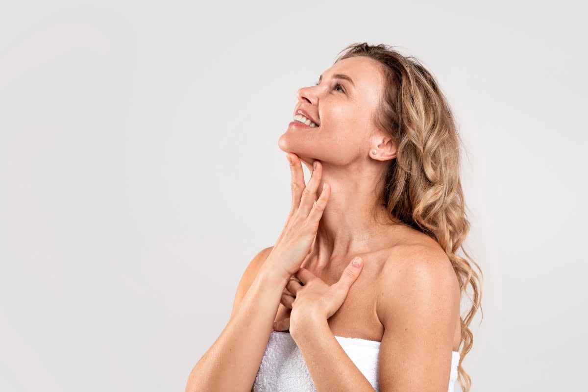 A woman with light hair wrapped in a white towel touches her neck and chin while looking upwards, contemplating the benefits of skin fillers against a plain background.