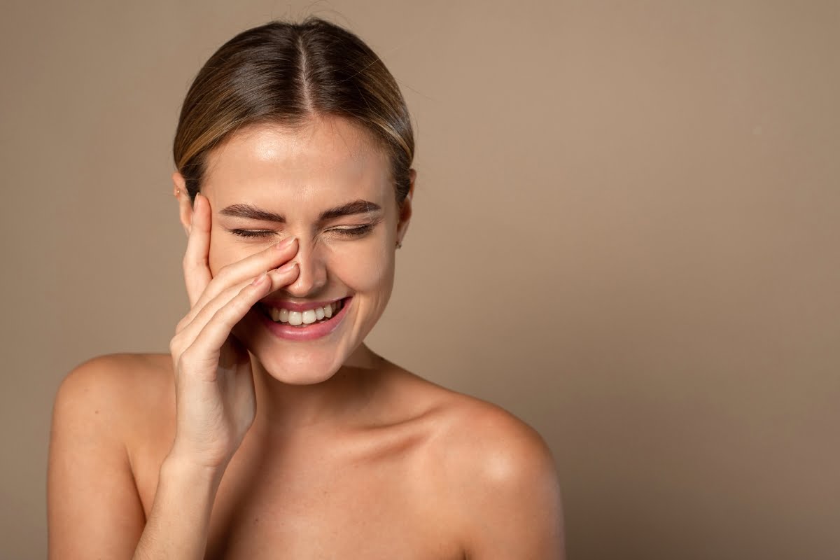 A woman with bare shoulders smiles with her eyes closed, touching her face with one hand, highlighting the smoothness achieved by skin fillers, against a plain background.