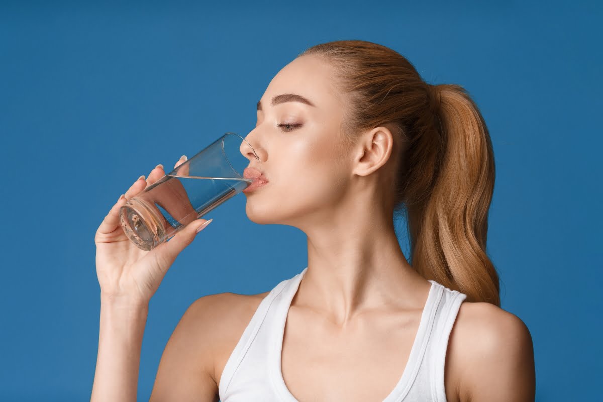 A person with long hair in a ponytail drinks water from a clear glass, showcasing microneedling benefits, against a blue background while wearing a white sleeveless shirt.