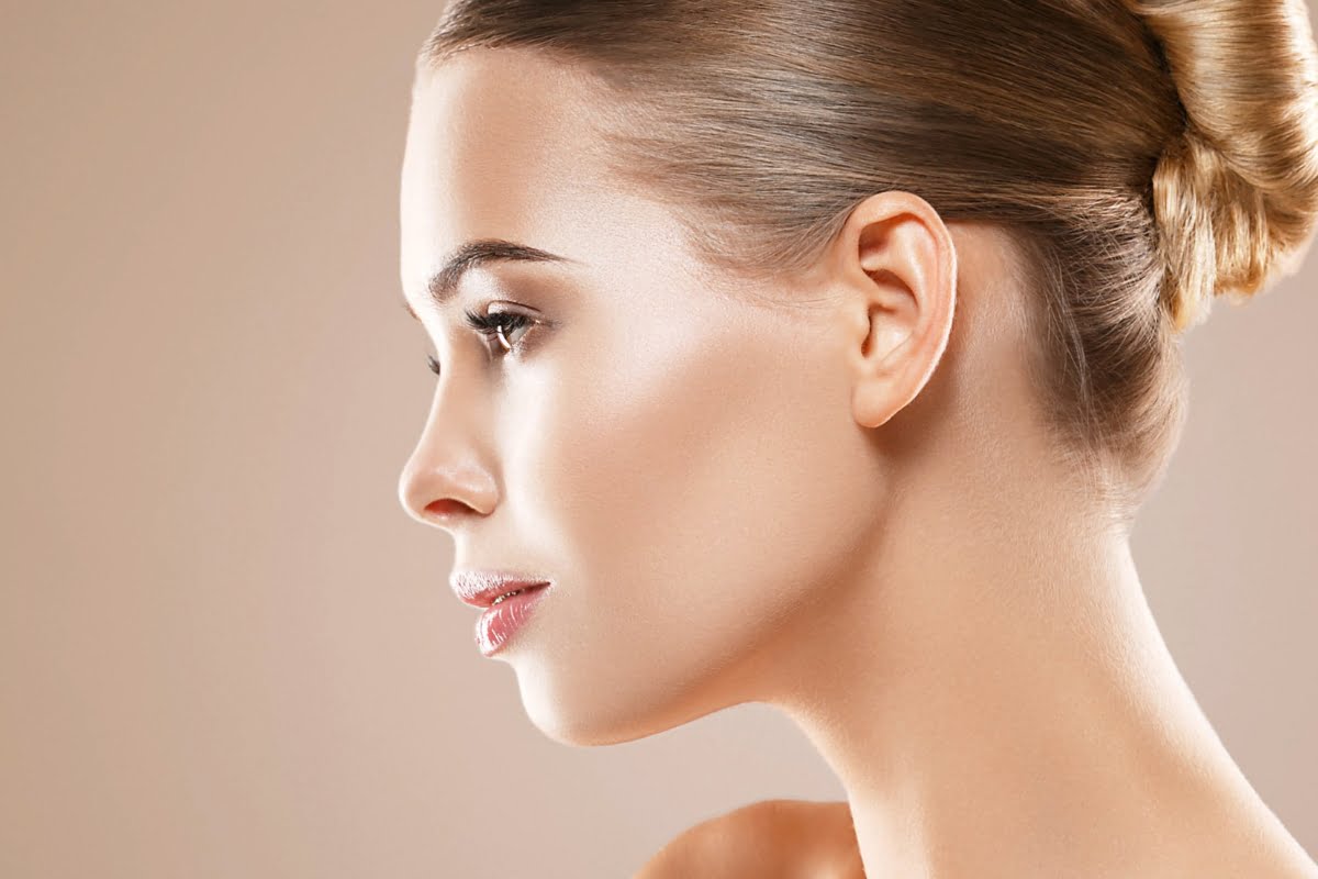 Profile view of a woman with smooth skin, thanks to microneedling benefits, and blonde hair neatly styled in a bun against a plain, beige background.