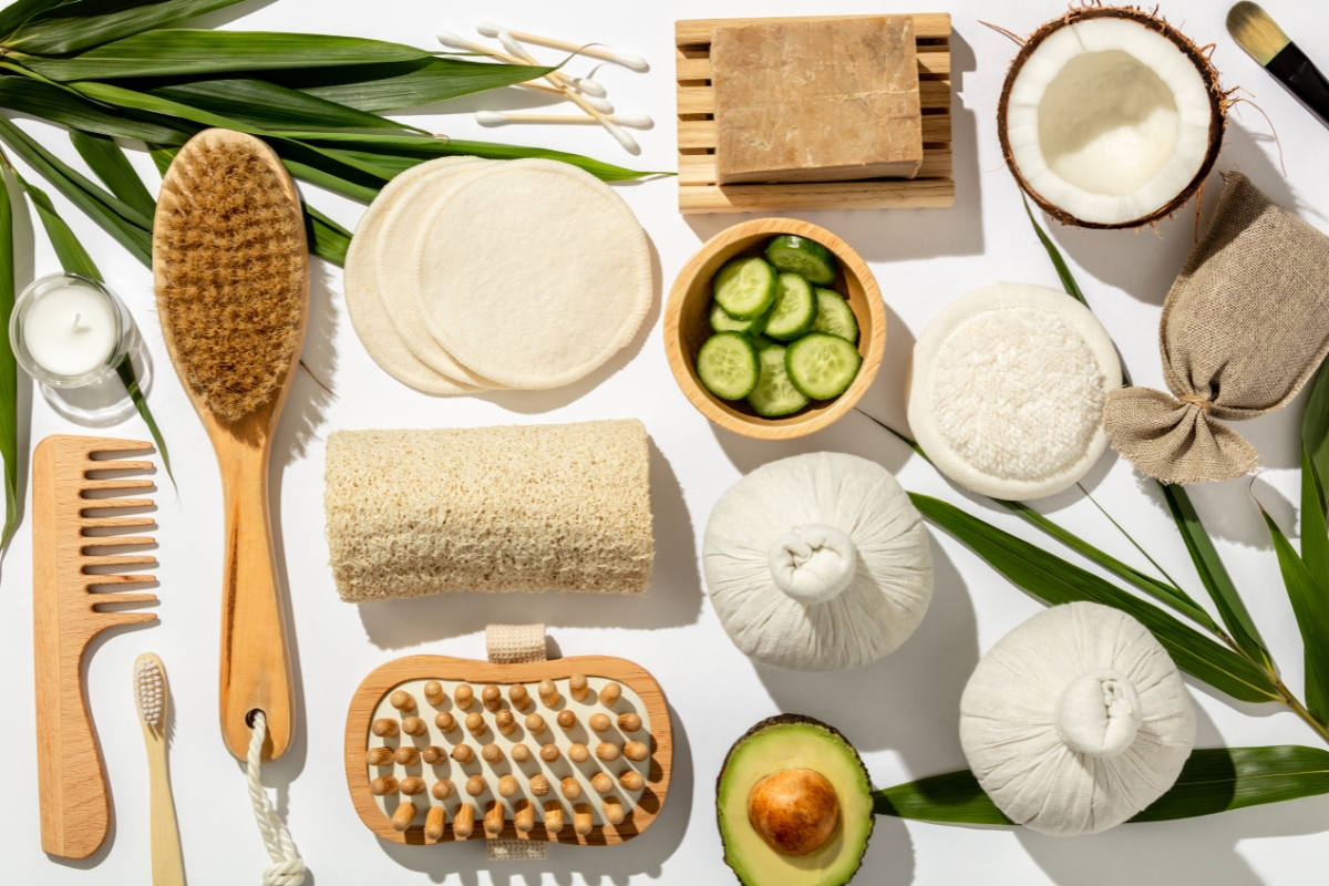 Various sustainable skin care and self-care items, such as a brush, soap, cotton pads, cucumbers, a sliced avocado, herbal bags, and leaves, arranged neatly on a light background.