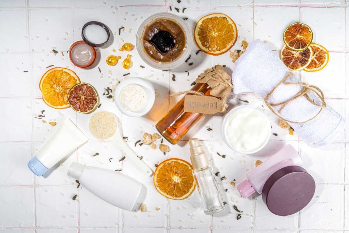 A flat lay of assorted sustainable skincare and beauty products, including creams, oils, lotions, dried orange slices, and spa accessories, arranged on a white tiled surface.