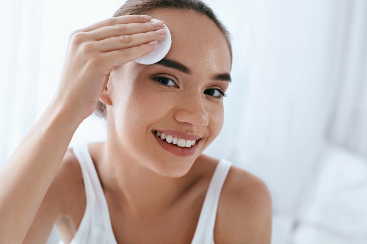 A woman in a white tank top smiles while using a cotton pad to clean her forehead, enjoying her sustainable skin care routine.