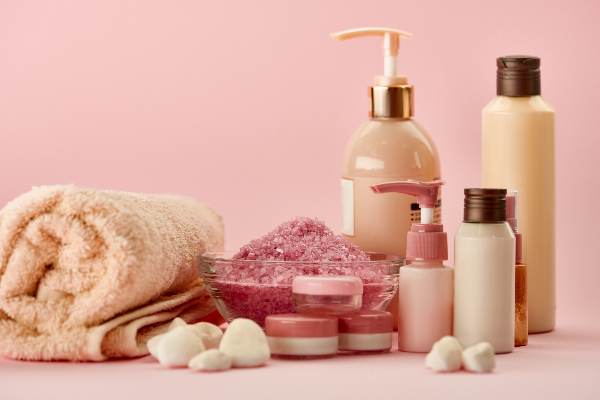 A collection of bath products, featuring a towel, bottles of lotion, jars of cream, and a bowl of pink bath salts—all arranged on a pink surface—highlights our commitment to sustainable skin care.