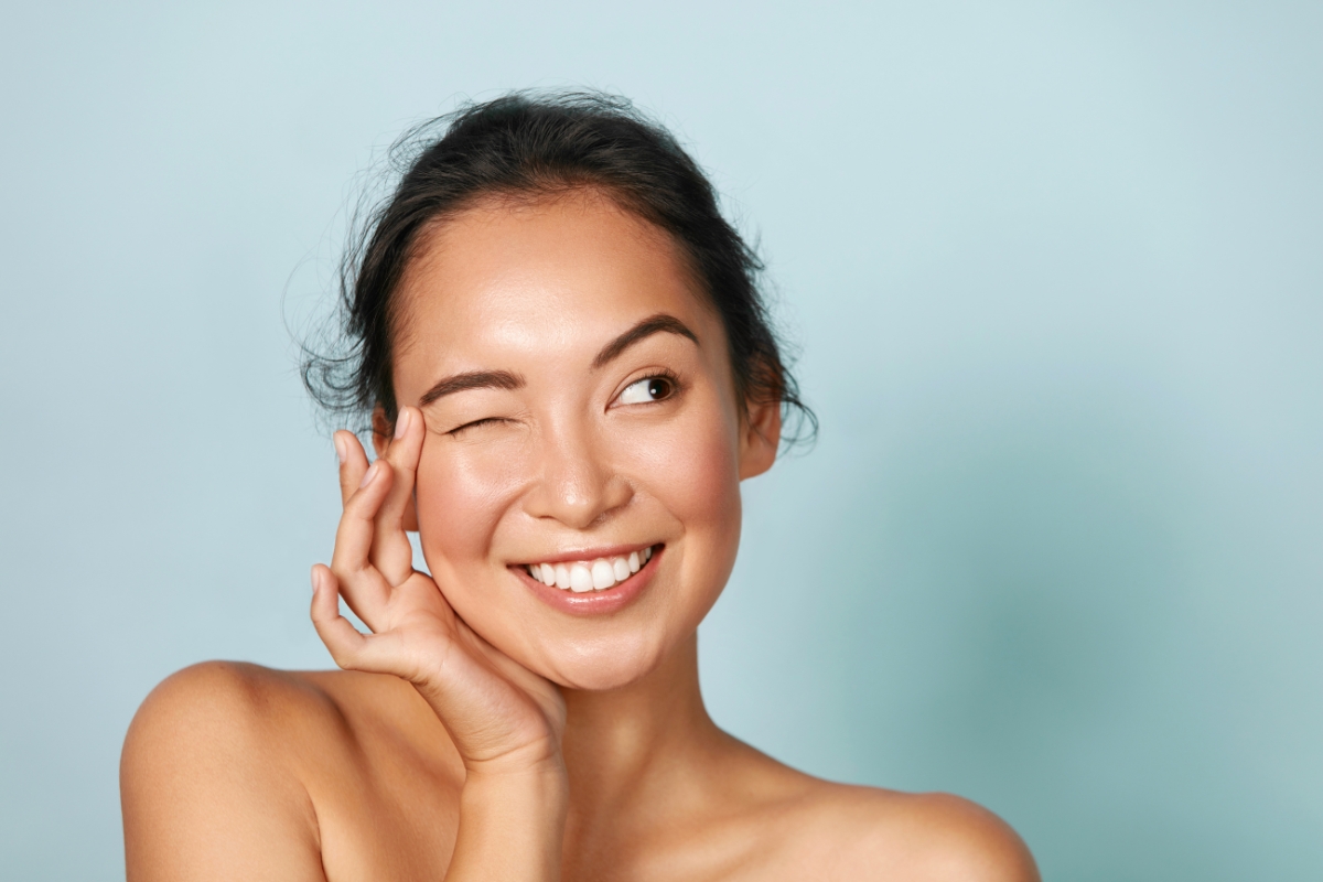 A woman with dark hair in a messy bun is smiling and winking. She is lightly touching the side of her face with her right hand, showcasing the radiance from her sustainable skin care routine. The background is solid light blue.