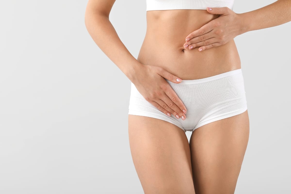 A woman in white underwear and a white top stands against a neutral background, with one hand on her stomach and the other on her lower abdomen, embodying the confidence and comfort often associated with non-invasive vaginal rejuvenation.