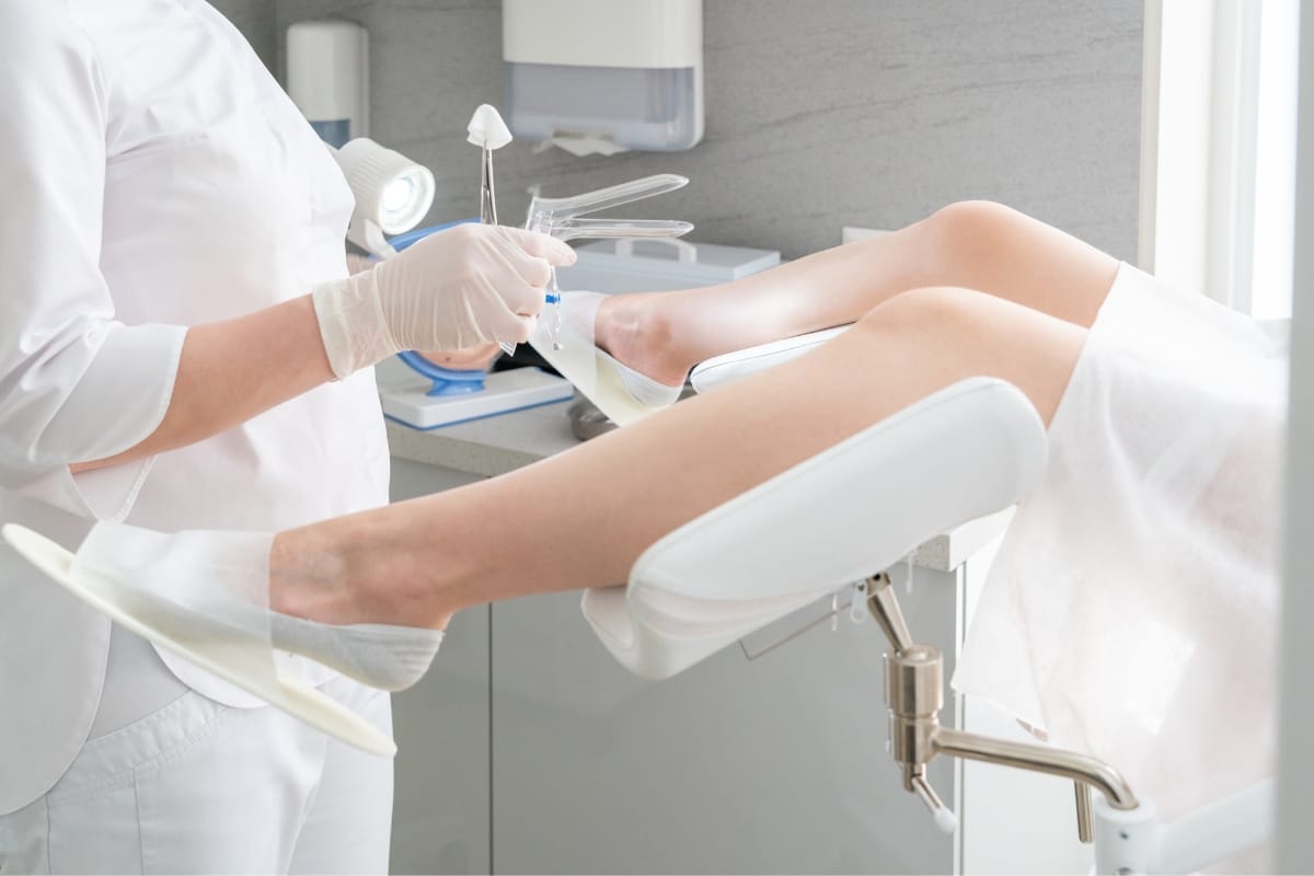 A gynecologist prepares medical tools for a pelvic examination while the patient lies on an examination table with legs in stirrups, discussing the option of non-invasive vaginal rejuvenation.