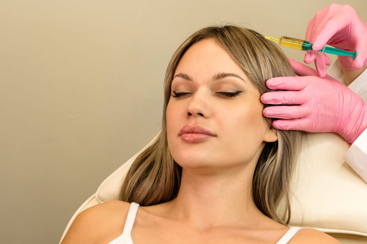 A woman with closed eyes is receiving a prp injection on the side of her forehead from a person wearing pink gloves and holding a syringe.